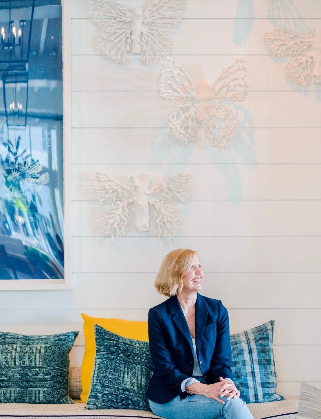 Lori Massicot sitting on a bench indoors, wearing a navy blazer and jeans, looking to the side beneath butterfly wall art, reflecting on living alcohol free in midlife.