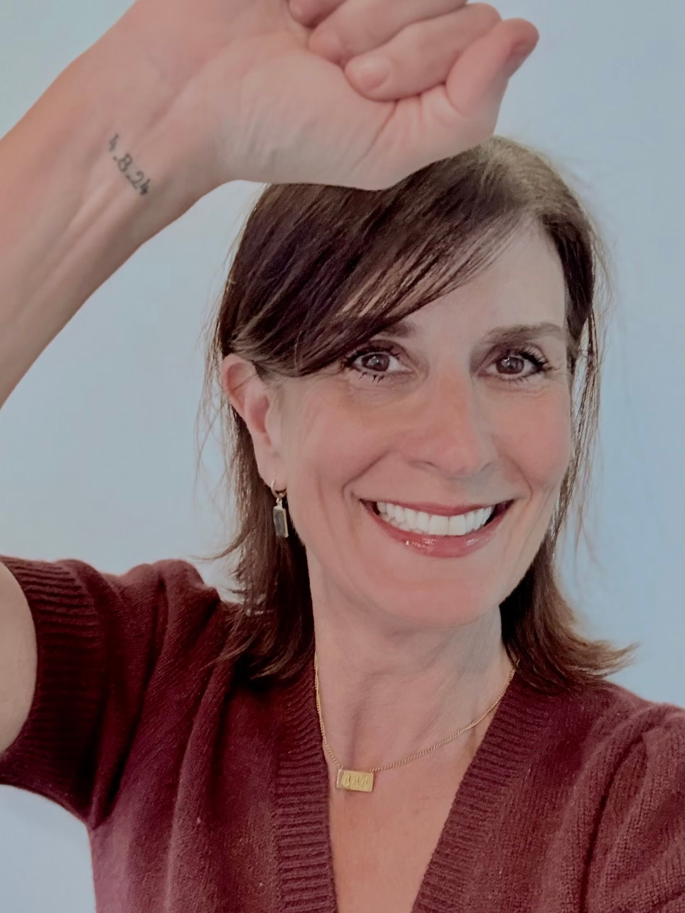 Smiling woman in a burgundy sweater raising her wrist toward the camera, showing a small “4.8.24” tattoo, against a light background.
