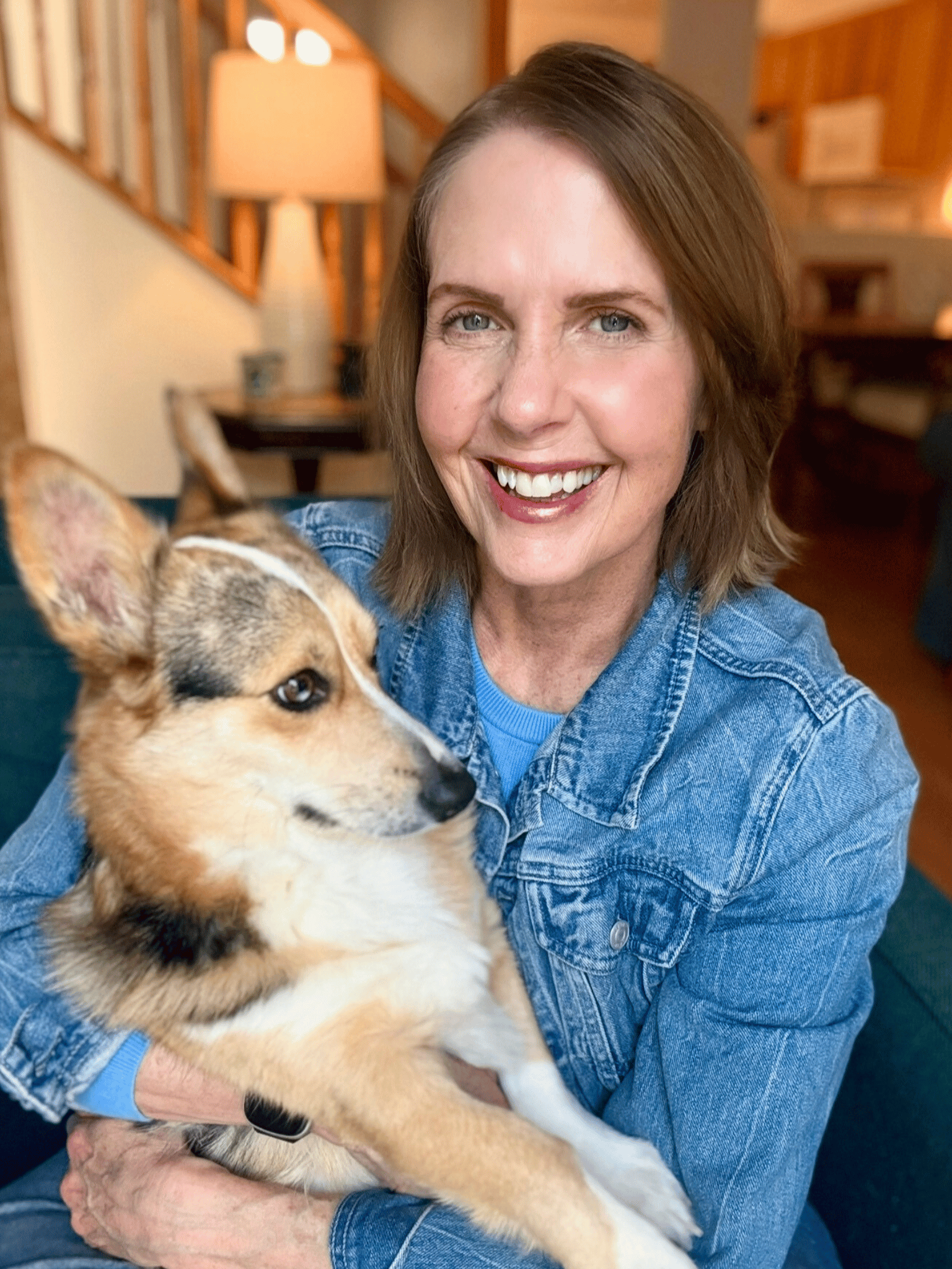Lori Massicot wearing a jean jacket, with dark blonde hair - looking at the camera smiling holding her dog a Cardigan Welsh Corgi, Franklin Blue.