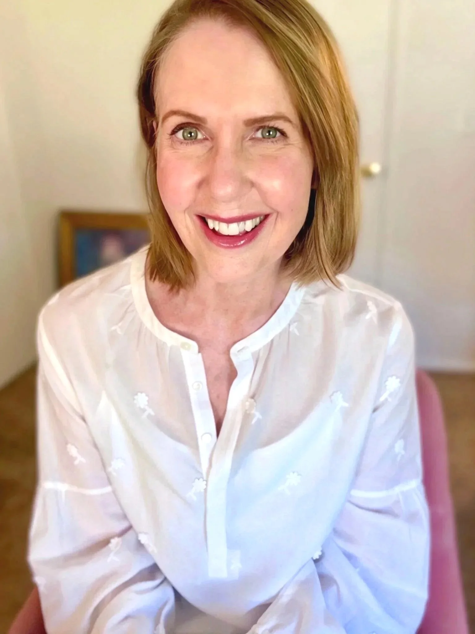Lori Massicot wearing a white blouse with flowers sitting in a pink chair looking at the camera smiling.