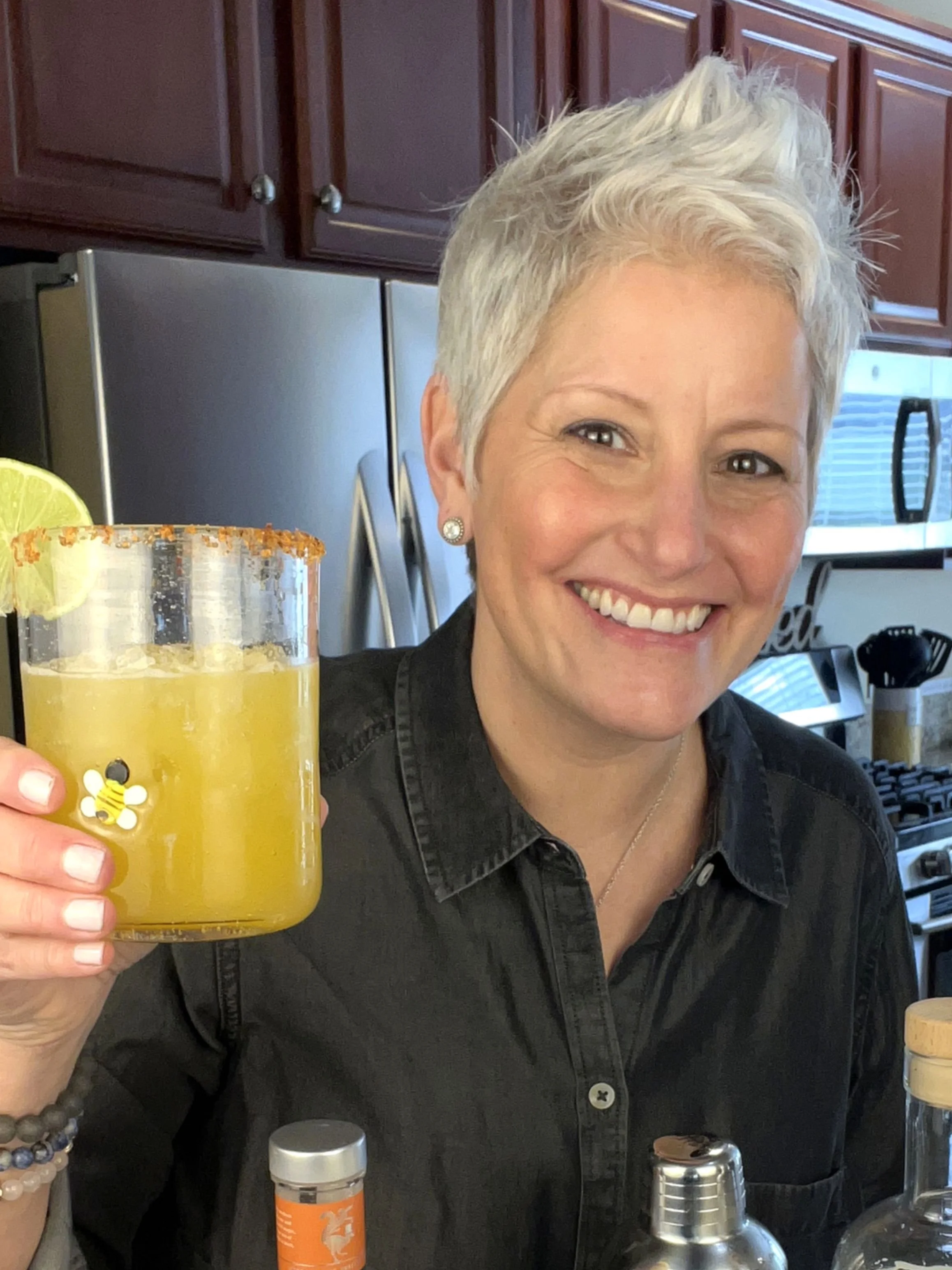 Smiling woman with short silver hair holds up a yellow mocktail with a lime garnish in a home kitchen, standing behind a counter with bar tools and bottles.