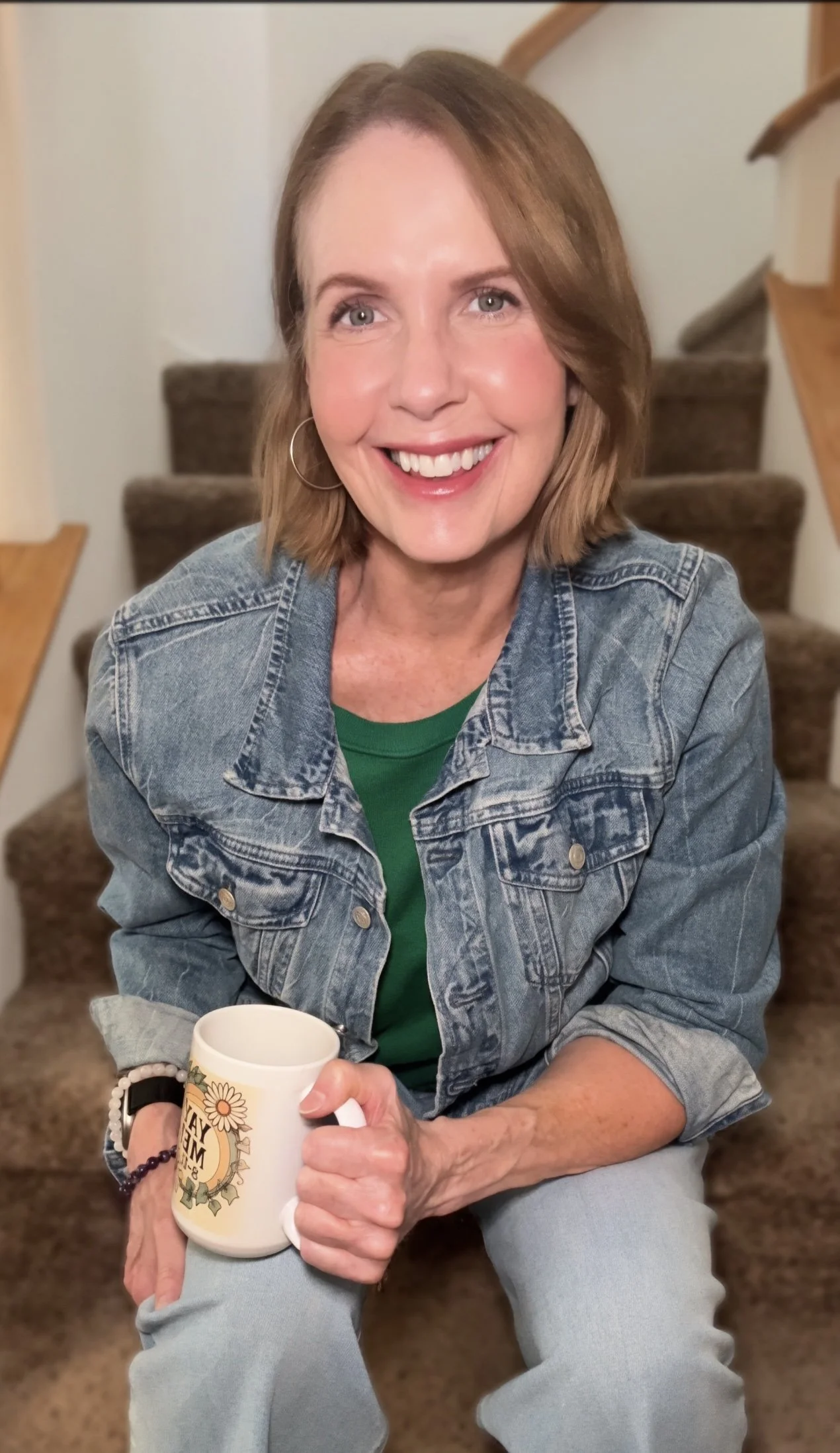 Lori Massicot sitting on the stairs smiling wearing a jean jacket and green sweater, blue jeans and holding a white coffee cup.