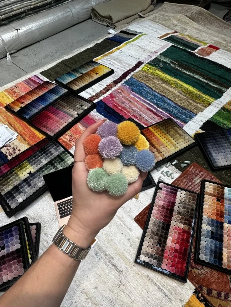 A person holding colorful fabric swatches and pom-poms in a workspace with fabric and textile samples spread out on a table.