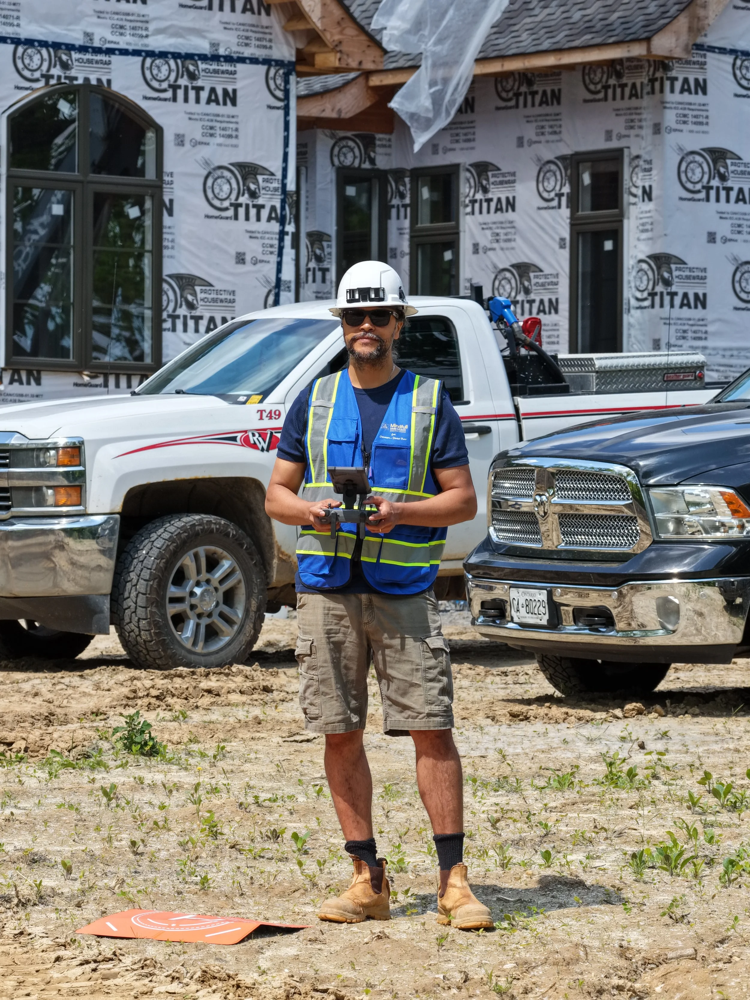 Construction worker standing on a dirt site holding a remote control. He is wearing a safety helmet, sunglasses, a blue safety vest, shorts, and boots. Behind him are two trucks and a house under construction wrapped in protective housewrap.