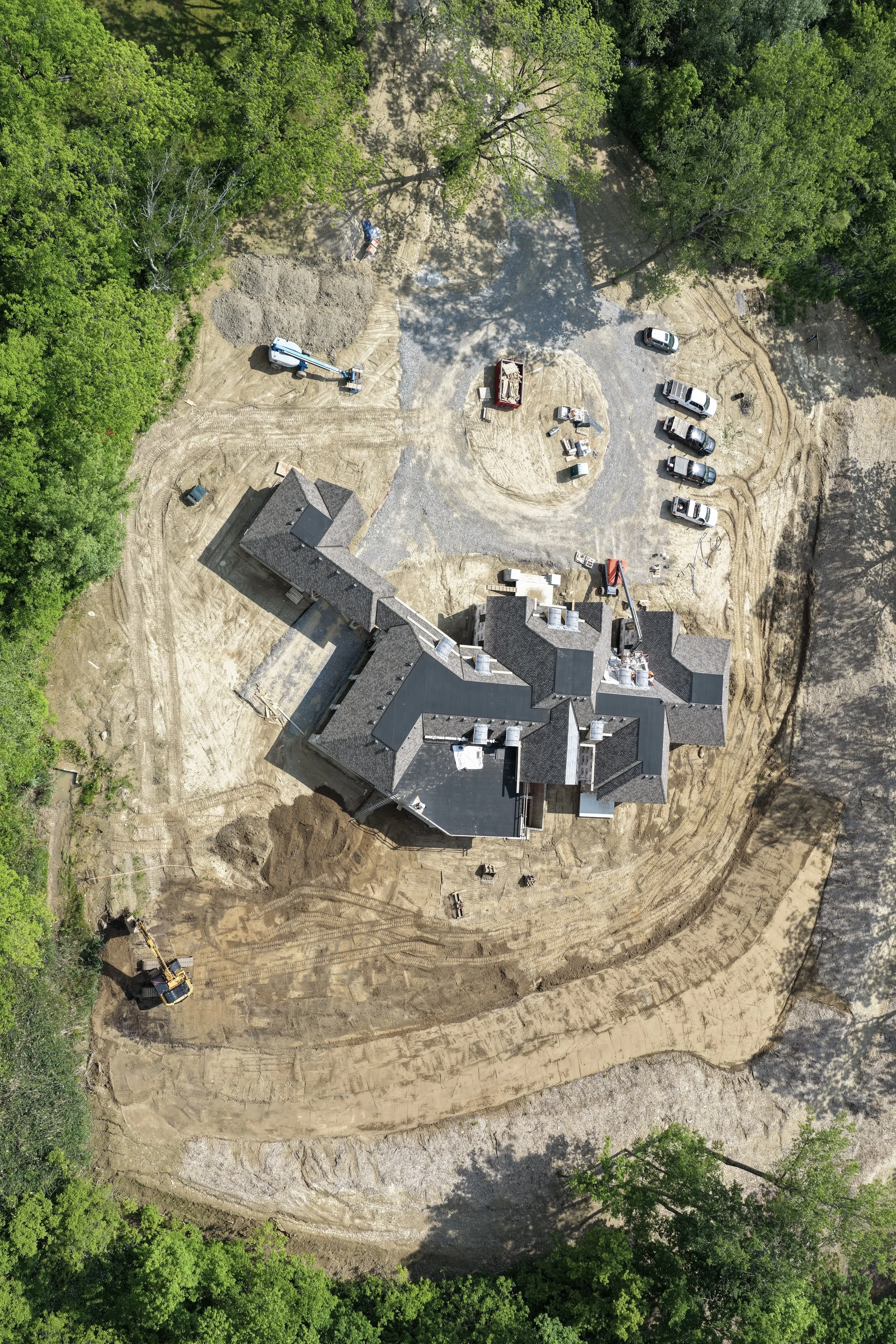 An aerial view of a house under construction surrounded by trees. Construction vehicles and materials are scattered around, and the ground is being excavated.