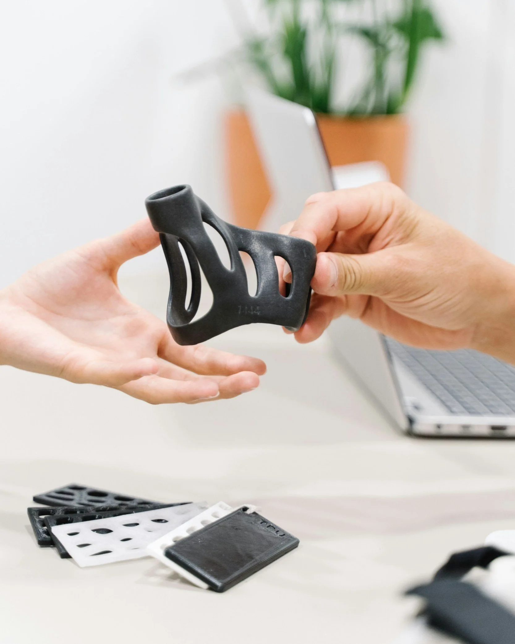 Person holding a black design prototype with another person's hand nearby in an office setting, with a laptop and plant in the background.