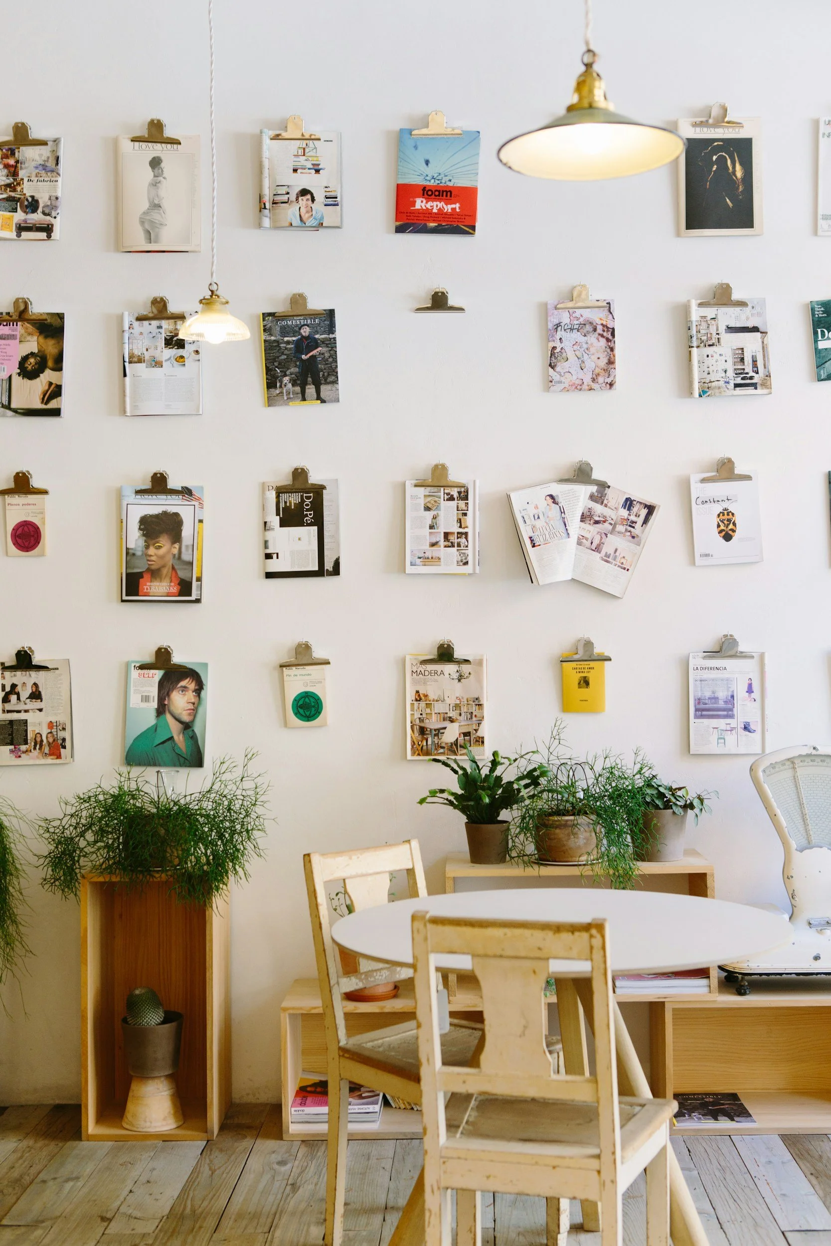 Interior scene with white wall decorated with magazines and artwork on clipboards, a white circular table, wooden chairs, and potted plants on wooden stands.