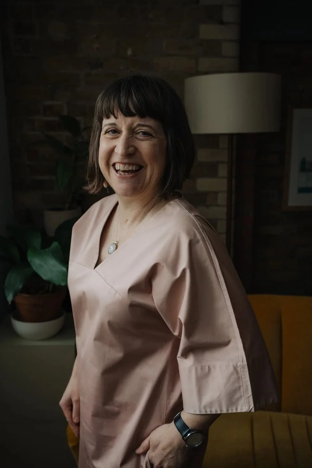 A woman smiling in a pink smock, standing indoors with a brick wall background, a potted plant, and a floor lamp.