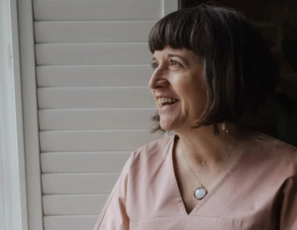 A woman with short dark hair and a fringe, smiling while looking out a window with light-colored horizontal blinds, wearing a blush-colored top and a necklace with a circular pendant.