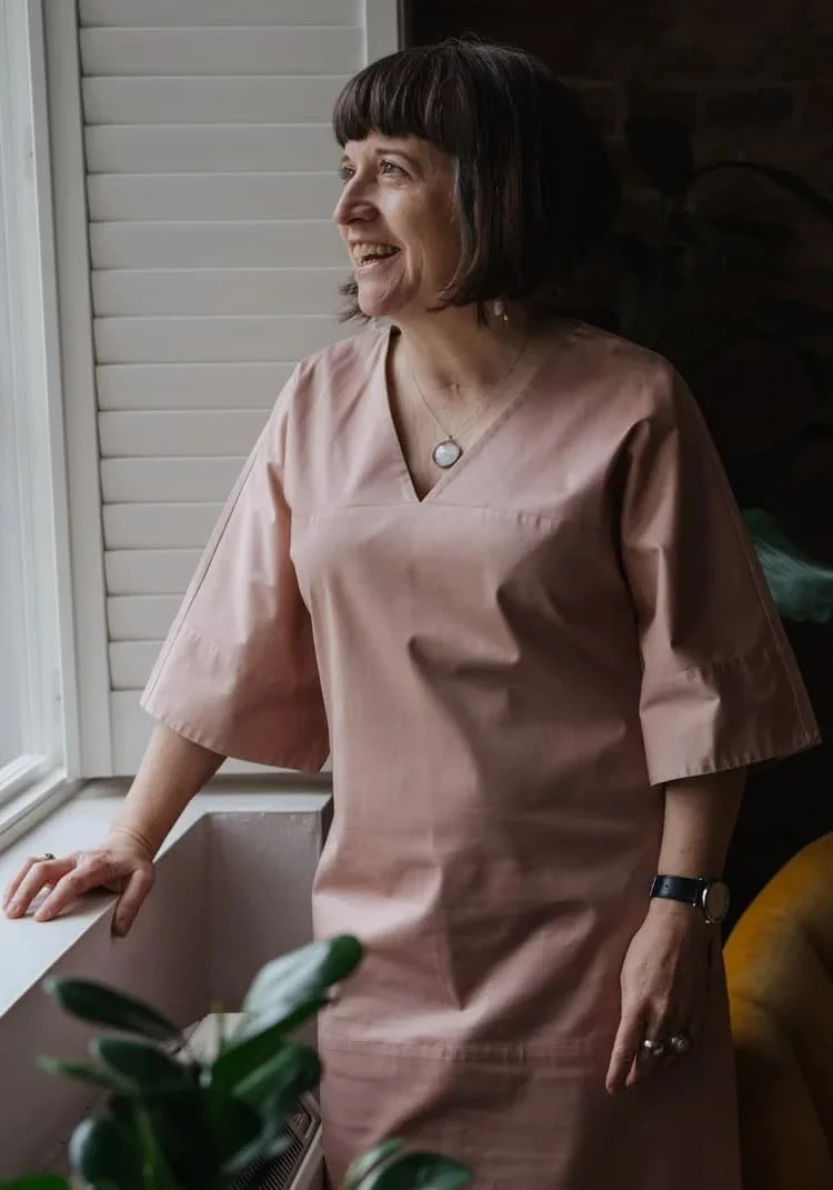 A woman with short dark hair, wearing a dusky pink dress, standing near a window and smiling while looking outside.