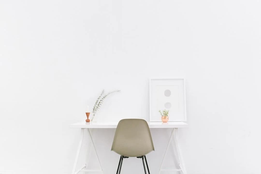 Minimalist white desk with a taupe chair, a small plant in a pink pot, a vase with a branch, and a framed artwork with two circles, set against a white wall.