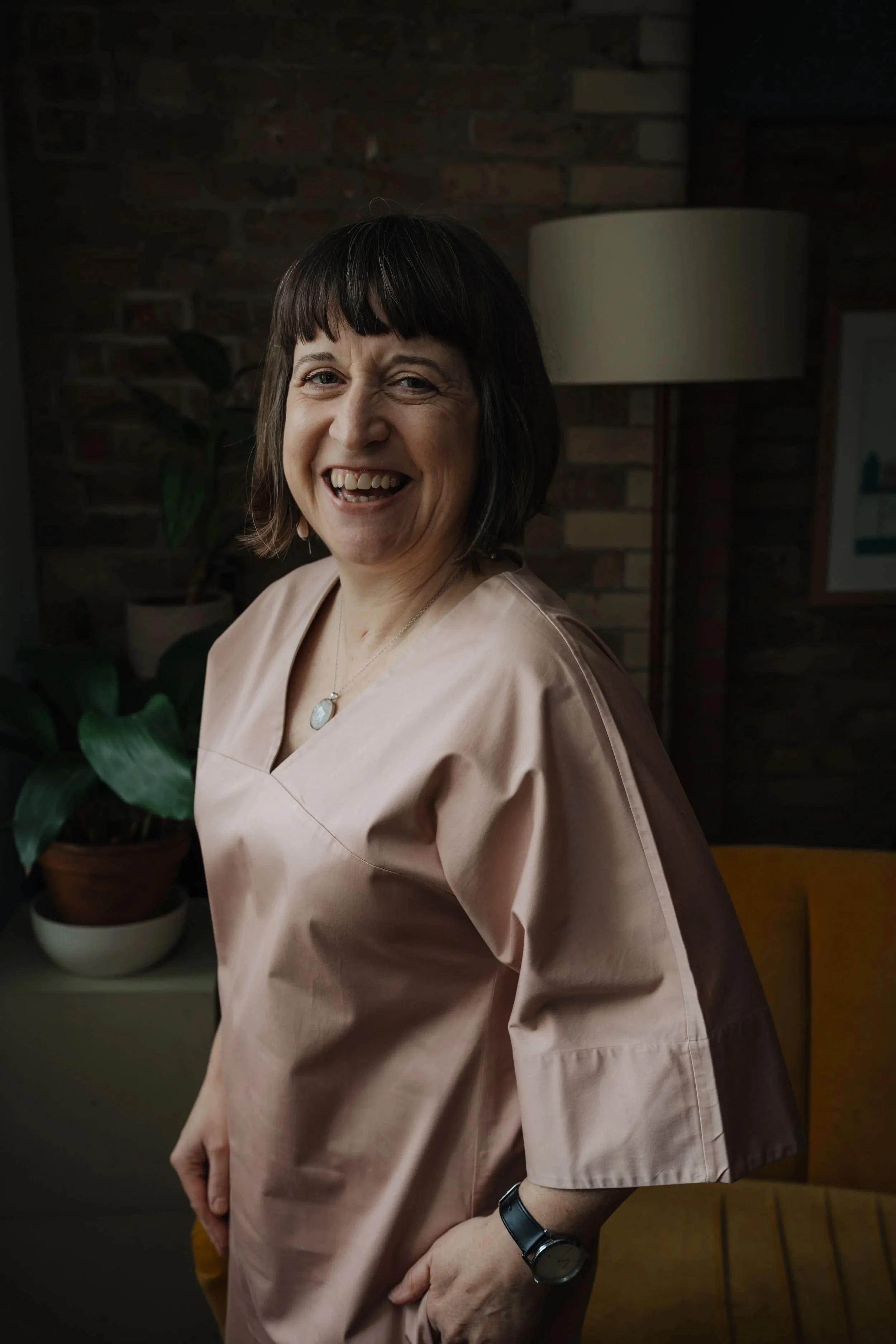A woman with short dark hair smiling, wearing a light pink scrub top, with a black watch on her left wrist, standing in a room with brick walls and houseplants in the background.