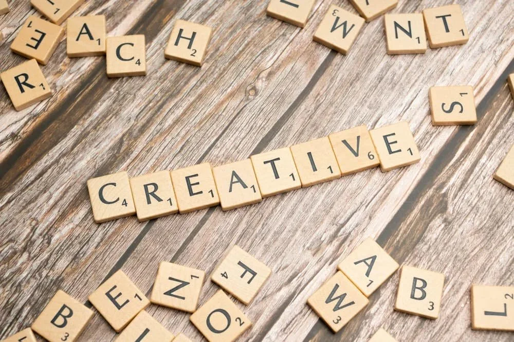Scrabble tiles spelling 'CREATIVE' placed on a wooden surface with scattered tiles around it.
