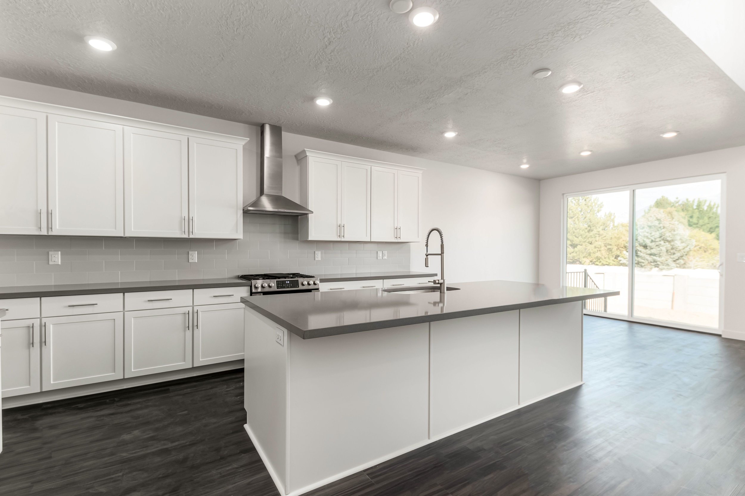 Modern kitchen with white cabinets, dark countertops, stainless steel appliances, and a large island. The room features recessed lighting, a high ceiling, and a view of the outdoors through large sliding glass doors.