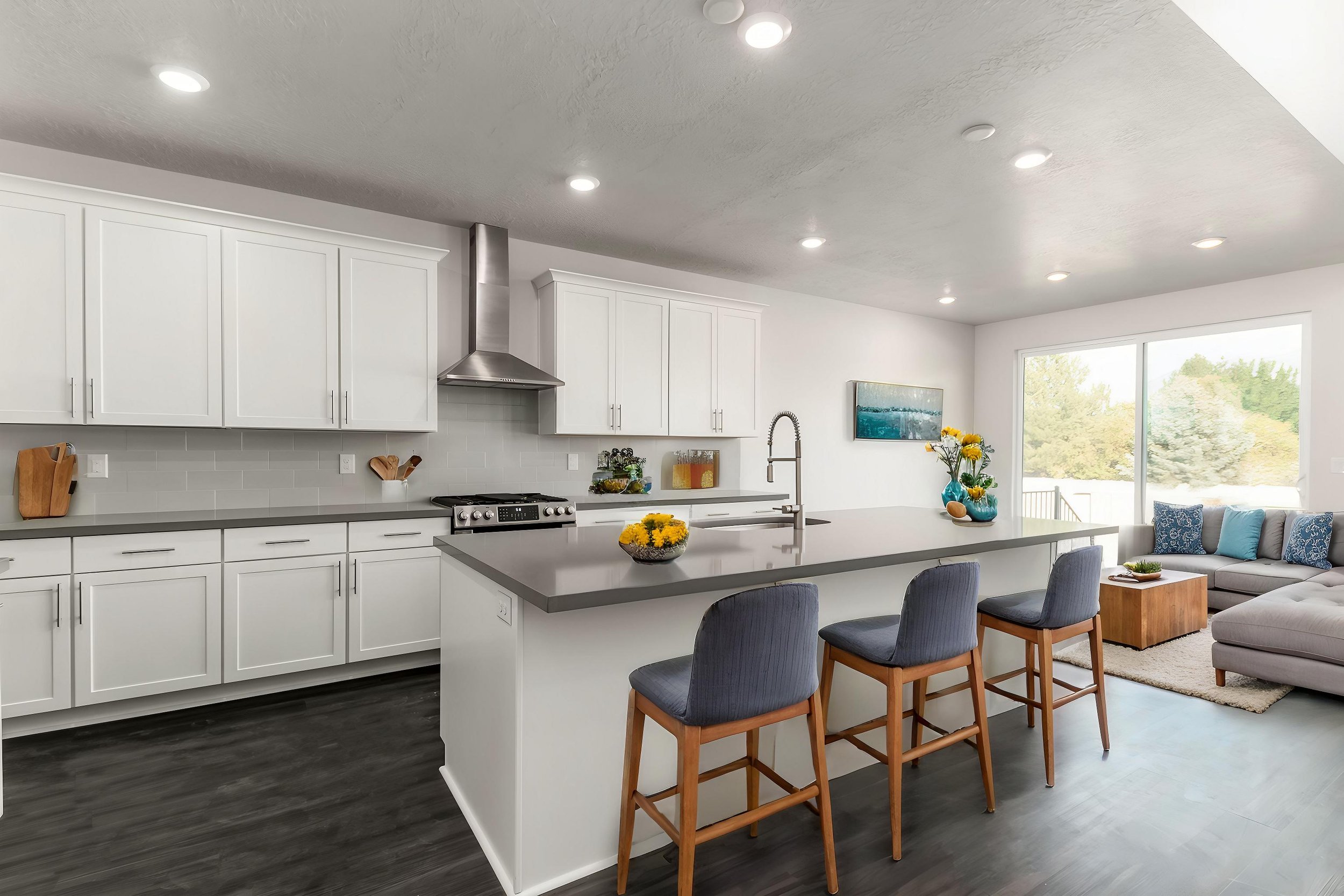 Modern kitchen interior with white cabinets, stainless steel appliances, and a gray countertop. Four bar stools at an island, flowers on countertop, and adjacent living area with a sofa and large window overlooking greenery.