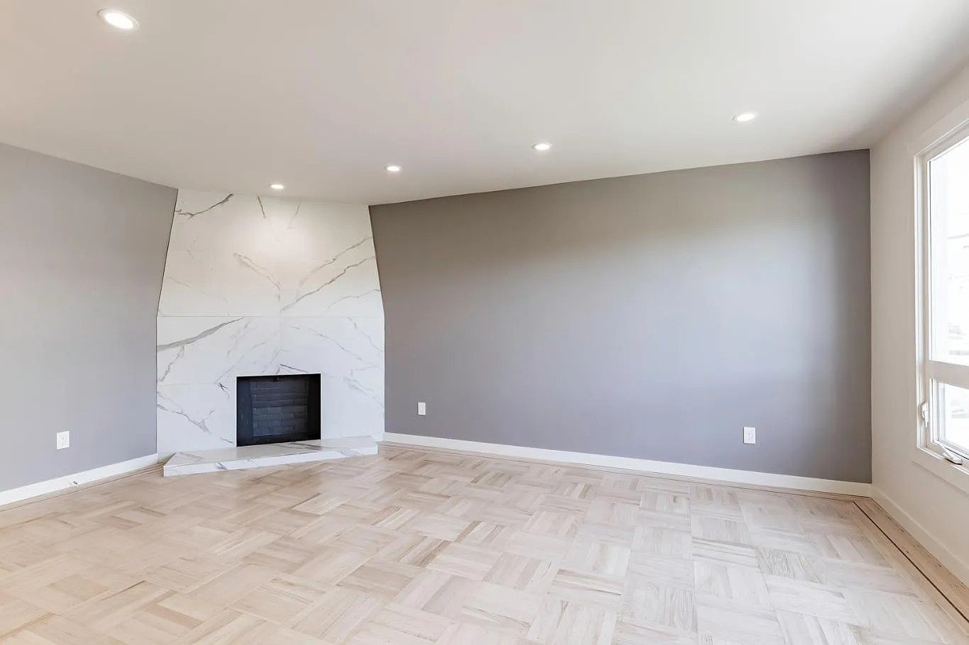 Empty room with light wood flooring, gray walls, recessed lighting, and a fireplace with a white marble surround.
