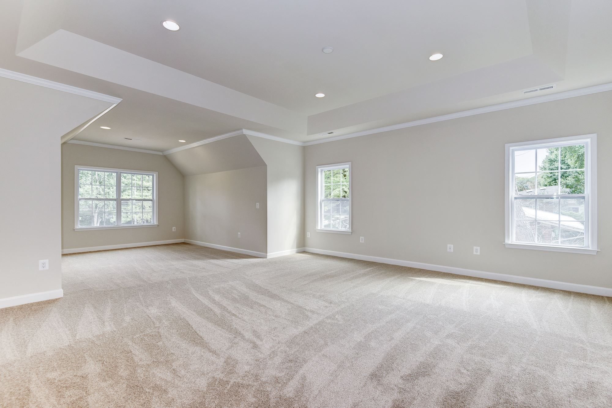Spacious empty room with beige carpet and light gray walls, featuring multiple windows and recessed lighting.
