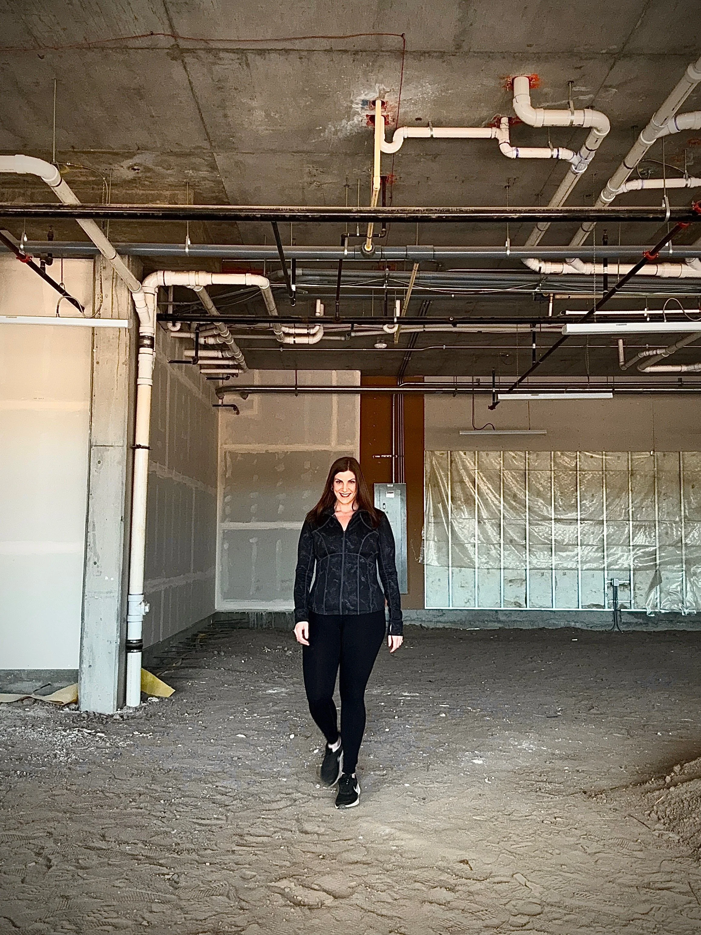 A woman with long dark hair wearing a black jacket and black pants jogging on a dirt surface inside an unfinished building with exposed pipes and concrete walls.