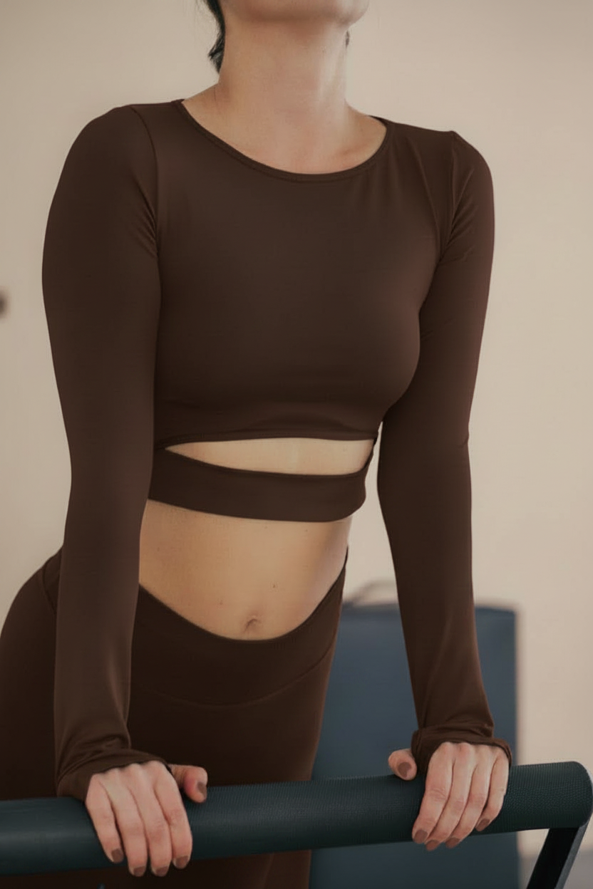 A woman wearing a brown long-sleeve crop top and matching pants, standing behind a black bar with her hands resting on it, in a neutral indoor setting.