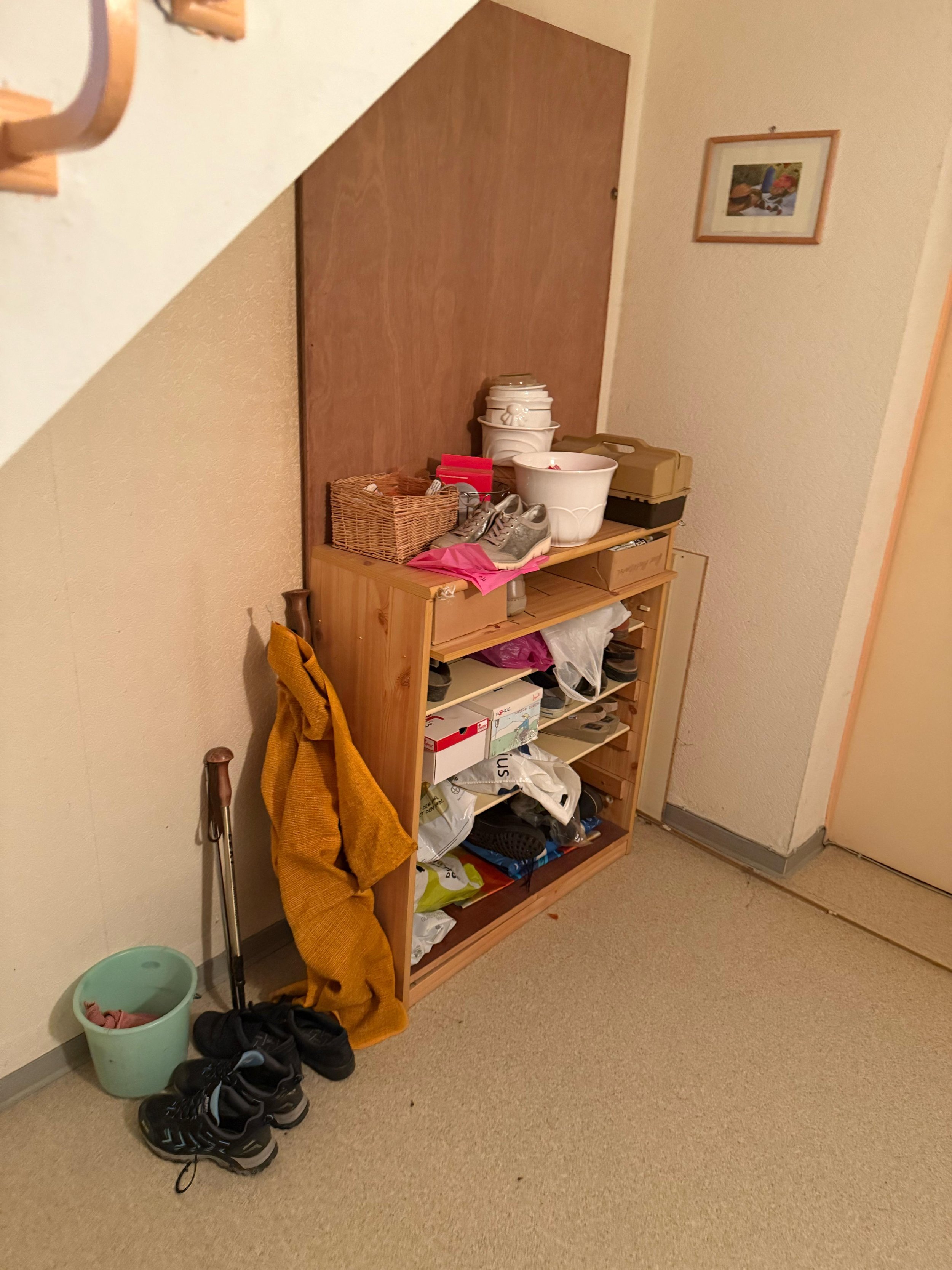 Flur mit Regal, auf dem diverse Gegenstände liegen, darunter Schuhe, Taschen und Kisten. Ein gelber Mantel hängt an der Seite. Ein kleiner Eimer steht auf dem Boden. An der Wand hängt ein gerahmtes Bild.