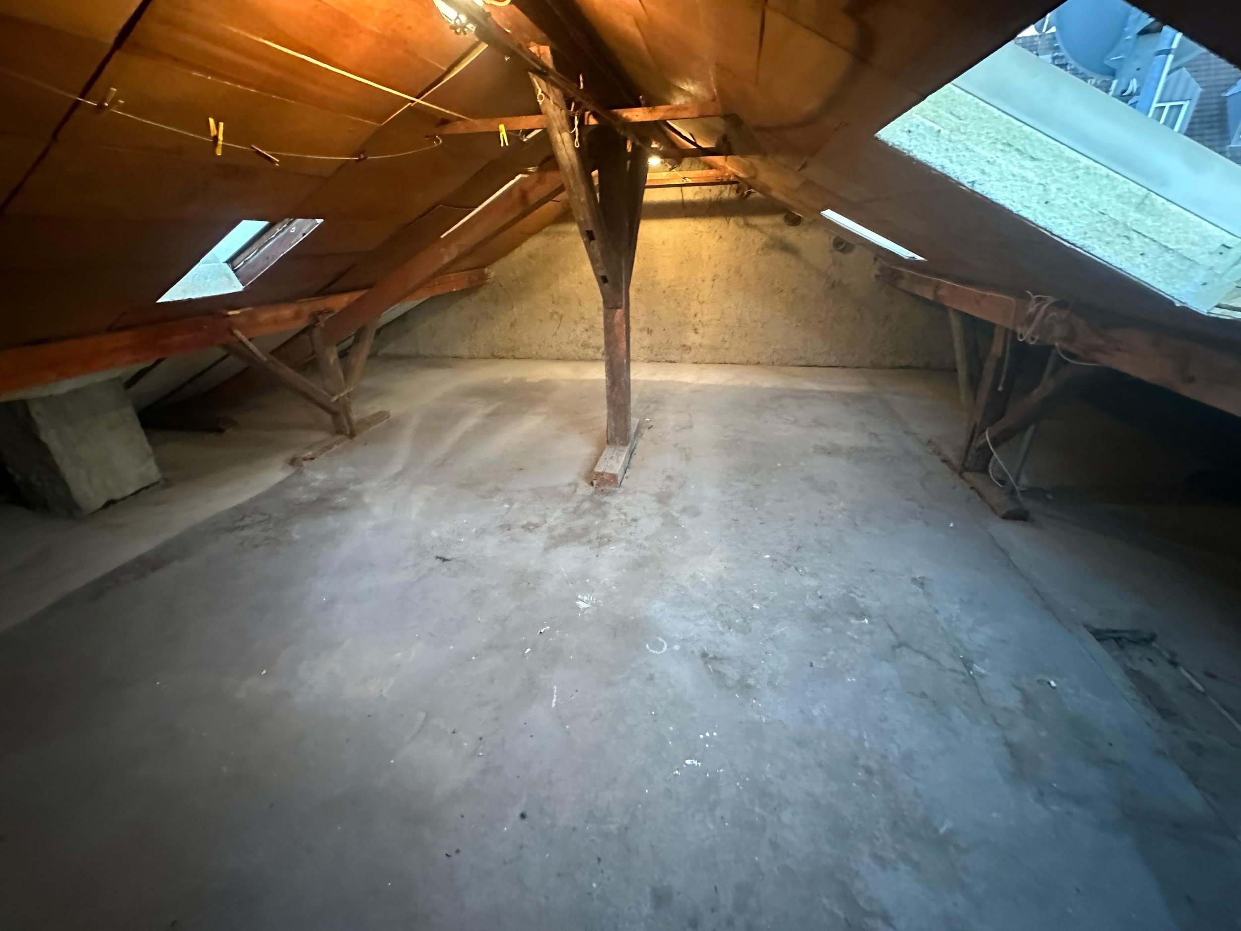Ein leerer Dachboden mit Holzbalken, zwei Dachfenstern und einer Glühbirne zur Beleuchtung. Der Boden ist staubig und aus Beton.
