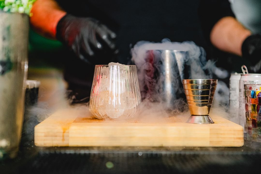 Bartender preparing a smoky cocktail with a glass of ice on a wooden board, surrounded by cocktail tools and vapor.