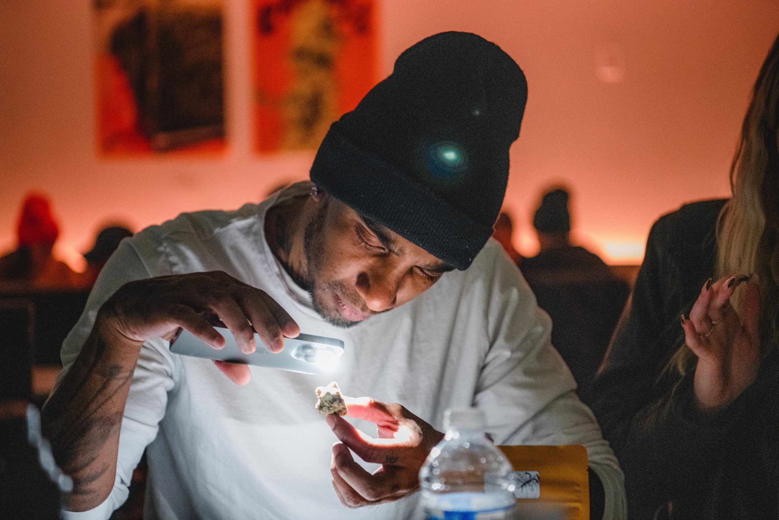 Person in white shirt examining an object under a light with a focused expression, seated at a table with a water bottle in the foreground. The background is softly lit with blurred figures and artwork on the wall.