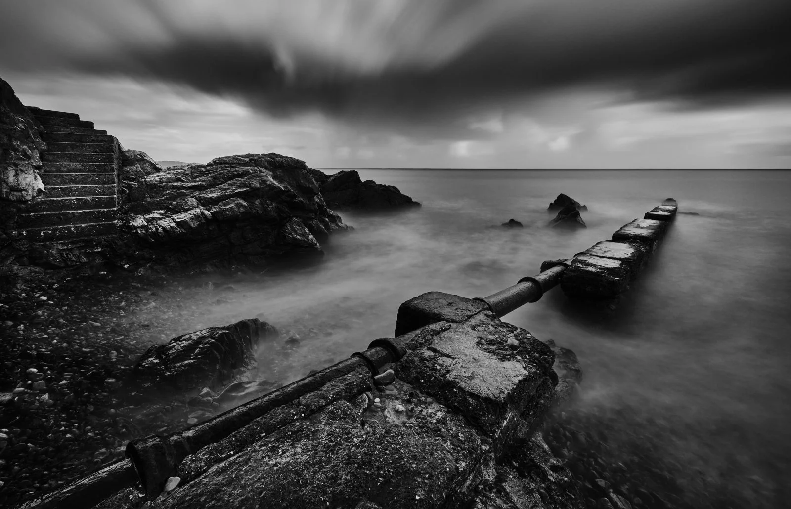 Black and white photo of a rocky seashore with steps on the left, a stone structure extending into the water on the right, and dramatic clouds overhead.