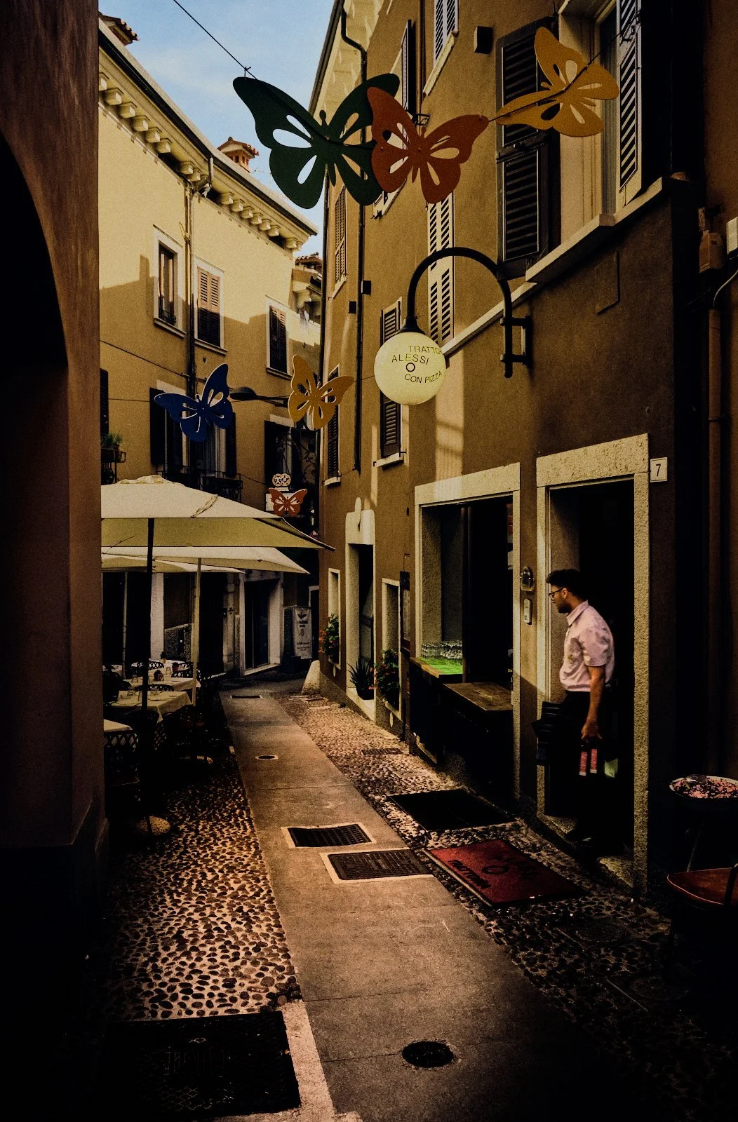 Decorative butterfly installations above a narrow street in Desenzano del Garda, a waiter standing in a sunlit trattoria doorway — Provincia Malinconica by Tom Imio
