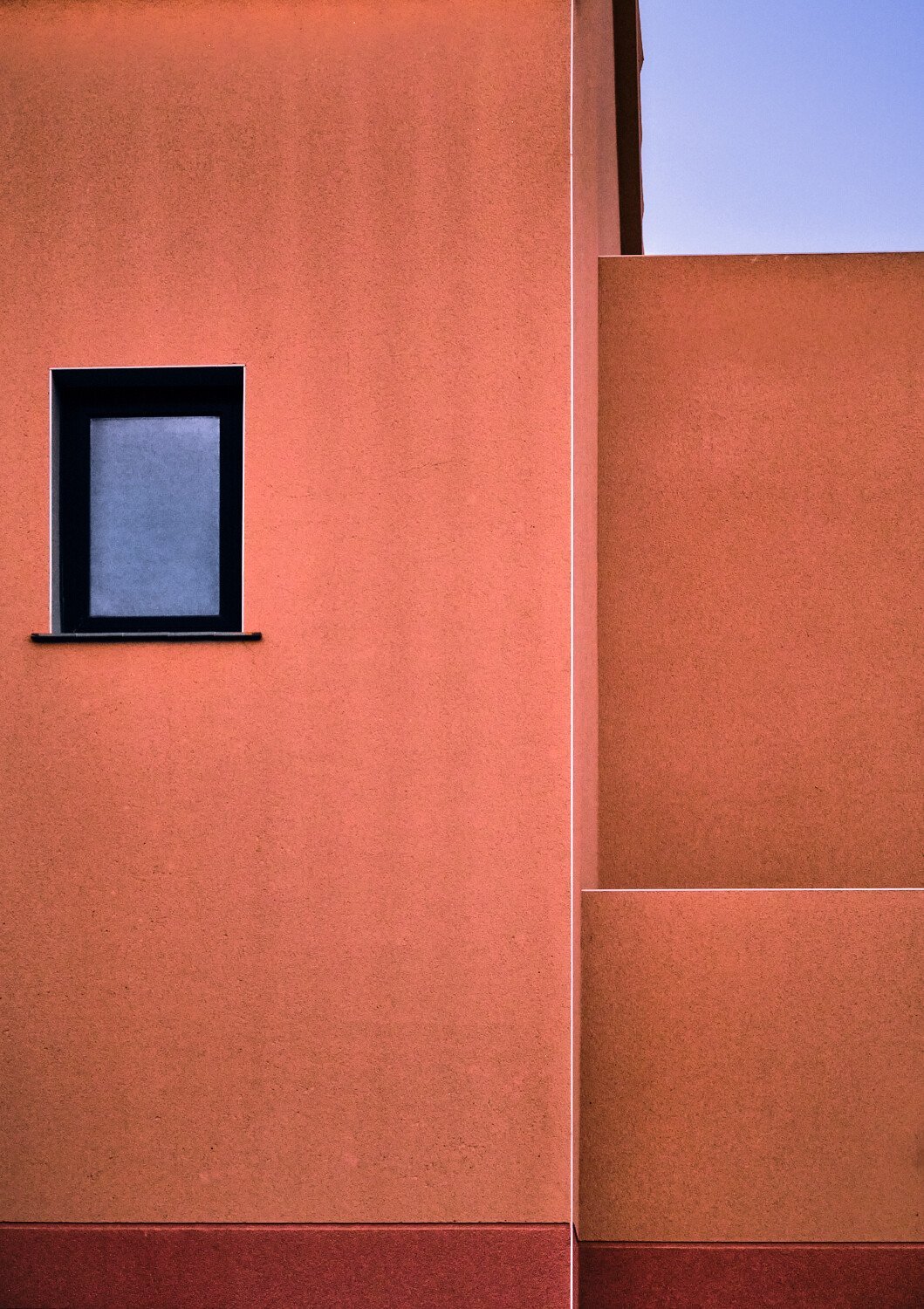 Close-up of a modern building's wall with a small black-framed window and minimalist architecture in warm terracotta colors.