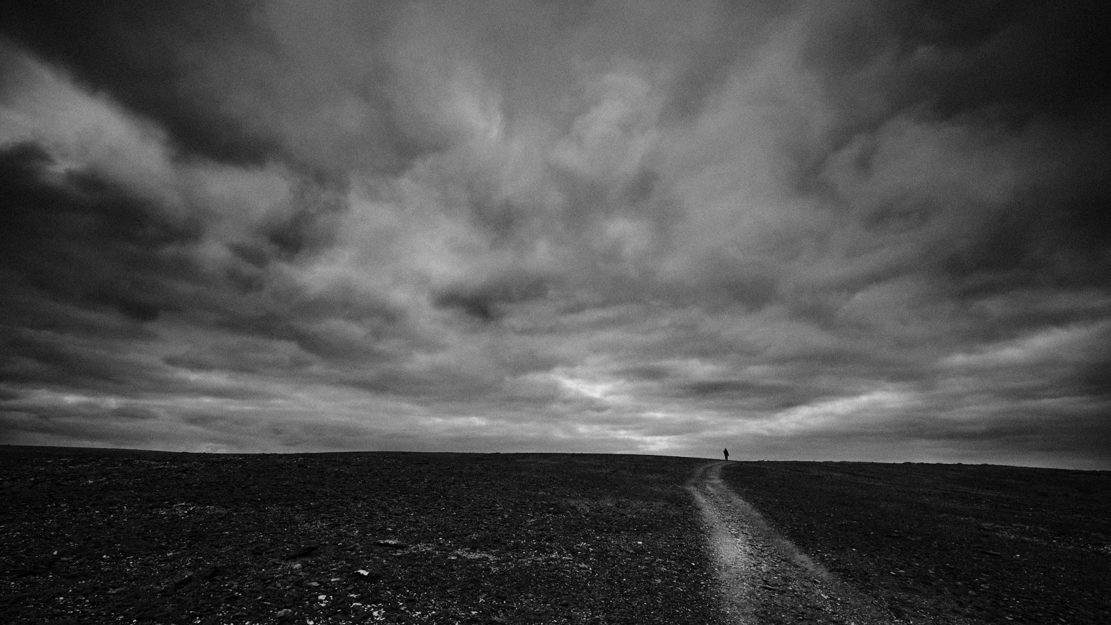 A solitary figure walking along a dirt path on a barren, dark landscape under a cloudy, overcast sky in black and white.