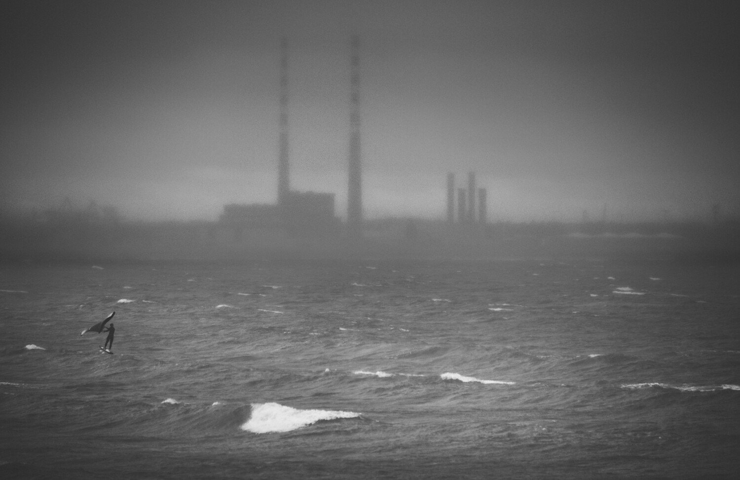 A person surfing on the ocean with a large industrial building with smokestacks in the distance, foggy and overcast.