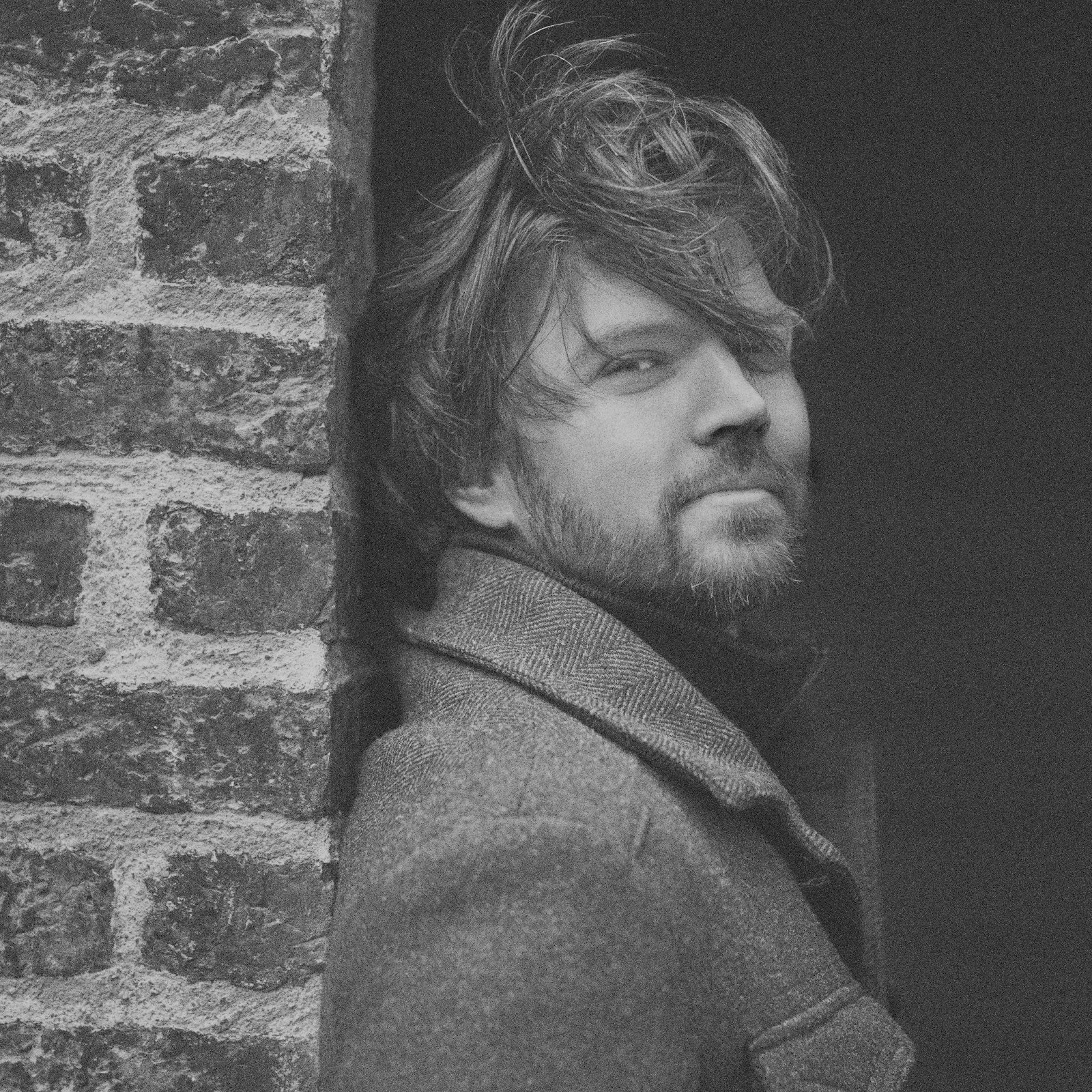 Black and white portrait of a bearded man in a herringbone coat leaning against a brick wall, half his face in shadow