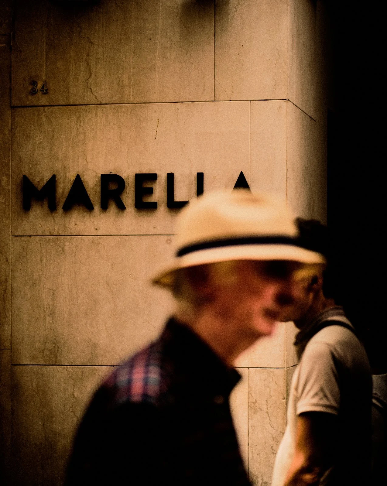 Blurred man in a panama hat passing a fashion boutique sign on a stone wall in Desenzano del Garda — Provincia Malinconica by Tom Imio