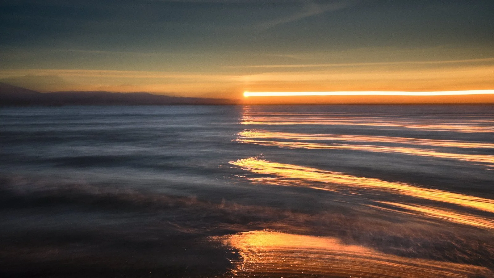 Sunset over calm ocean waters with reflections on the surface and a distant landmass on the horizon.