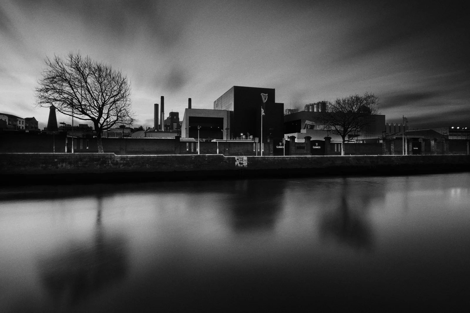 Black and white photo of an industrial building with two leafless trees in front, reflection of trees and building in water, cloudy sky at dusk.