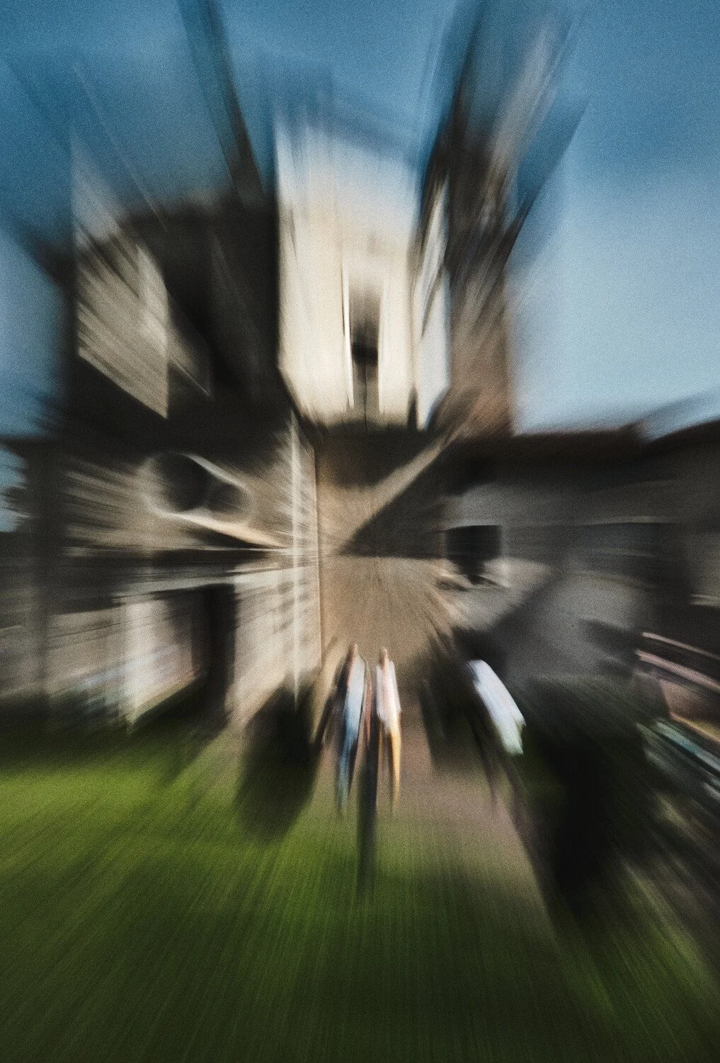Blurred photo of a city street with buildings on both sides, several pedestrians walking, and parked cars, taken from above.