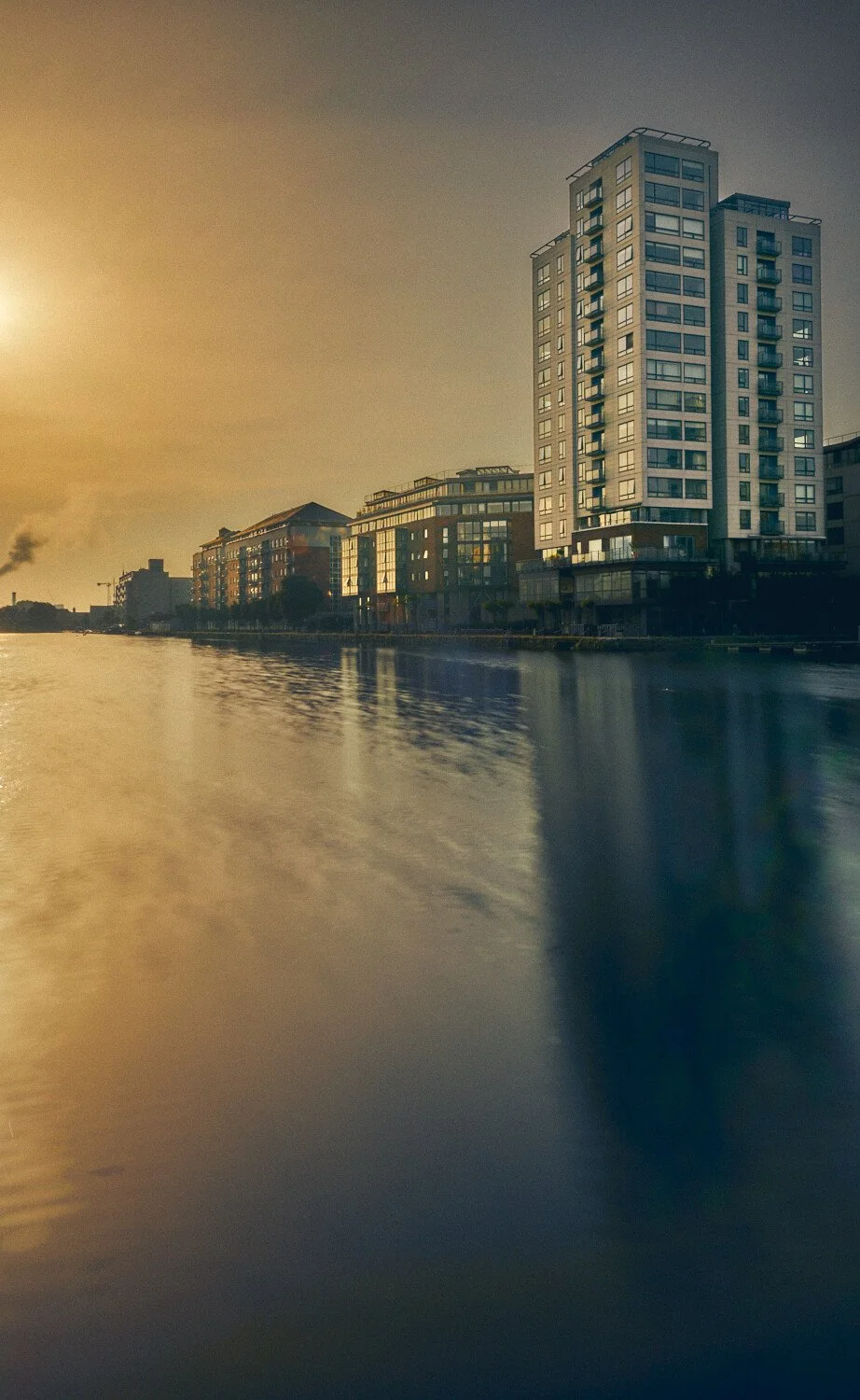 High-rise modern apartment buildings along a waterway during sunset, with reflections on the water and an industrial factory in the background.