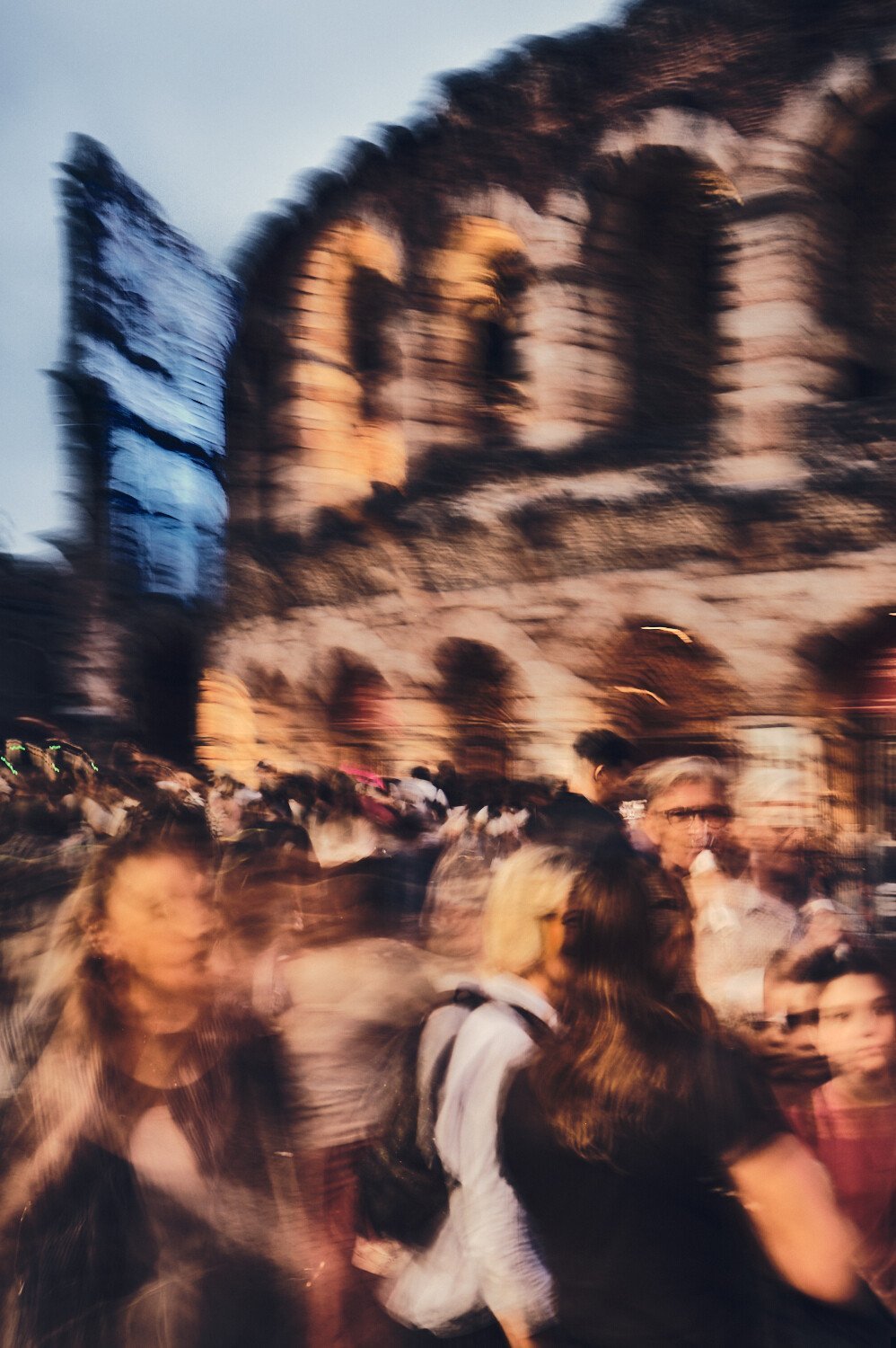 Crowd of people walking in front of a historic stone building with an arched structure, blurred due to motion, during dusk or evening.