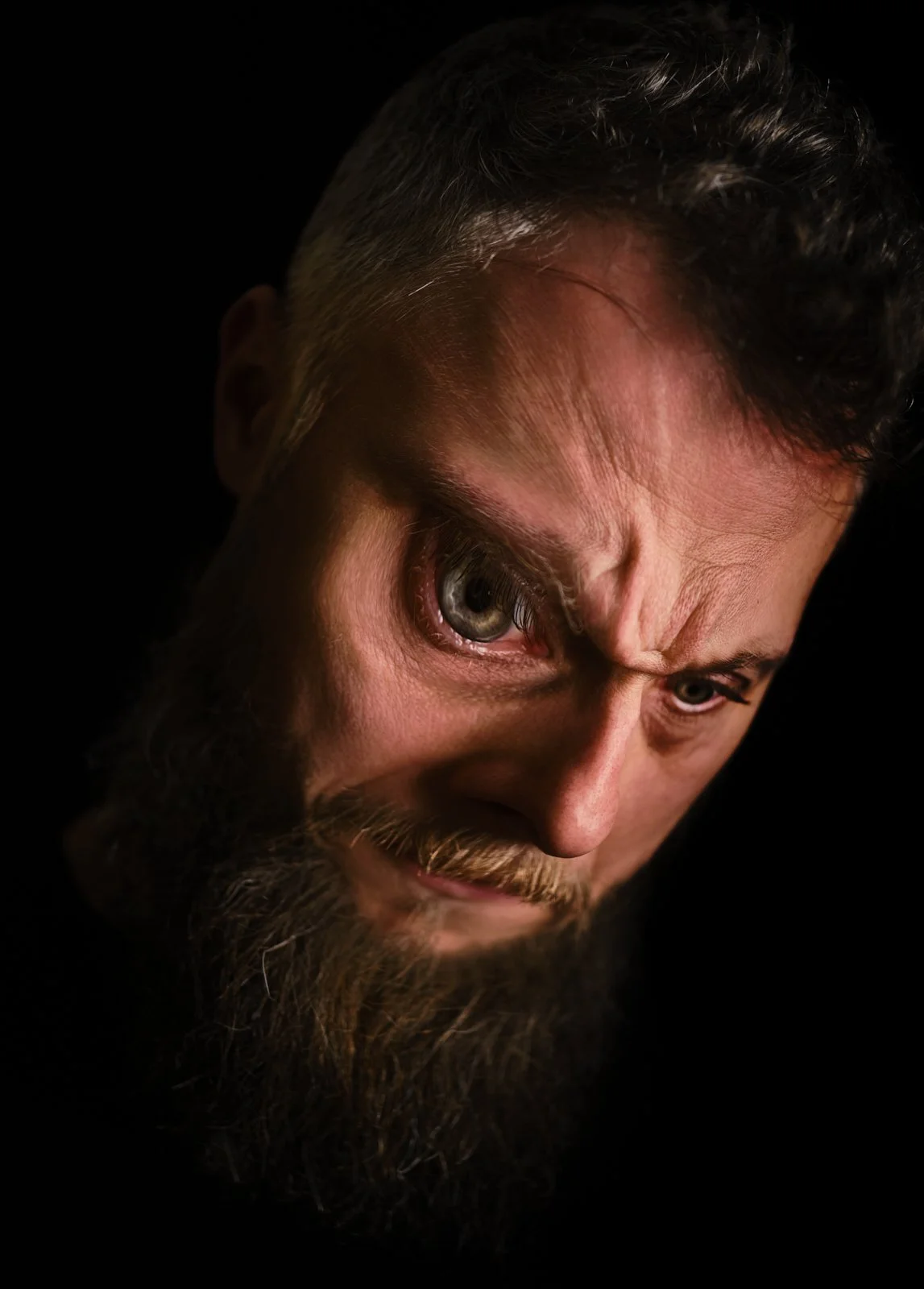 Close-up of a man's face with intense expression, beard, mustache, and gray hair, against a black background.