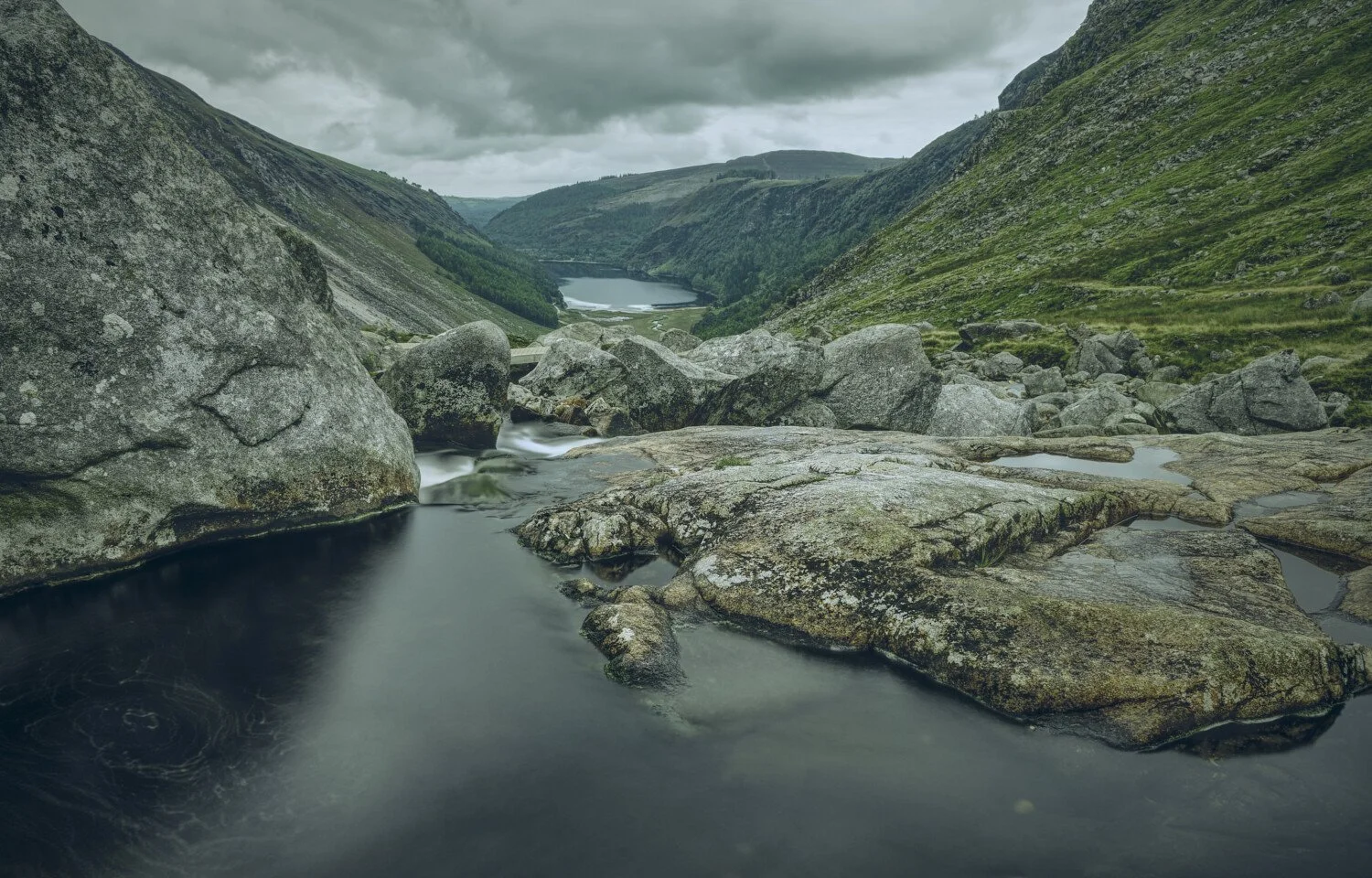 A rocky mountain landscape with a flowing stream and a distant lake surrounded by green hills under a cloudy sky.