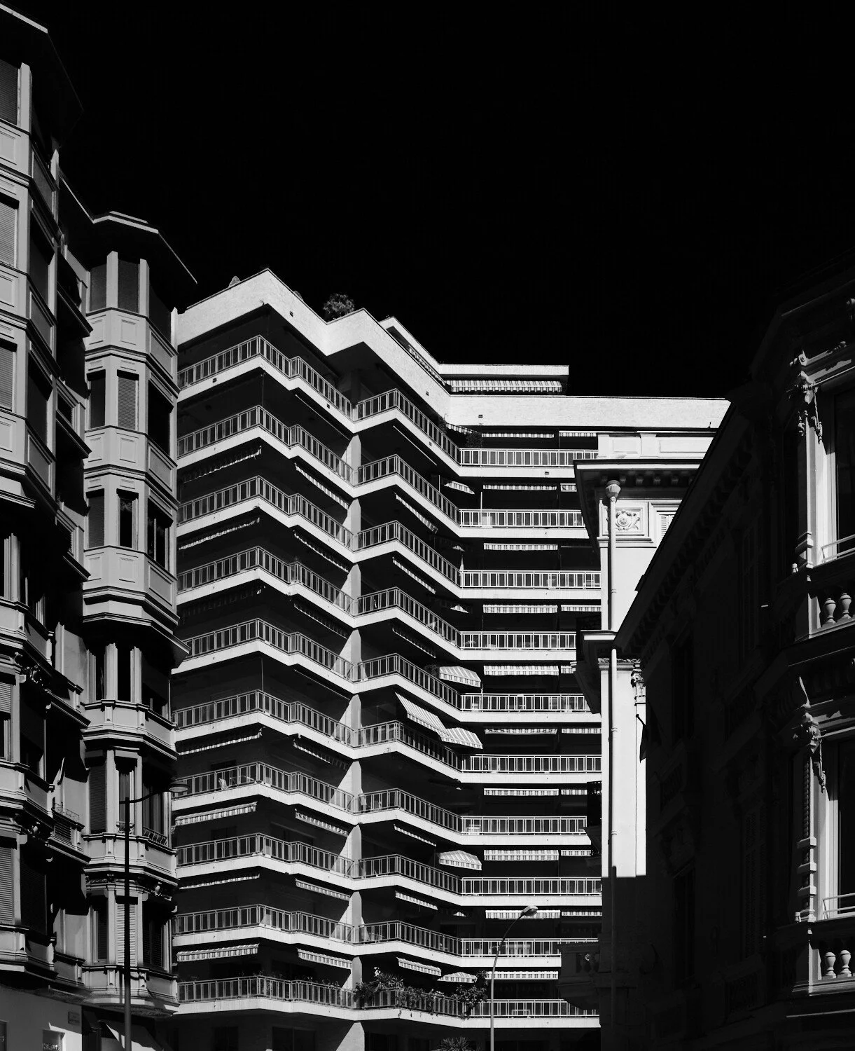 Black and white photo of modern high-rise apartment buildings with balconies, surrounded by older, ornate buildings with decorative facades and street lamps.