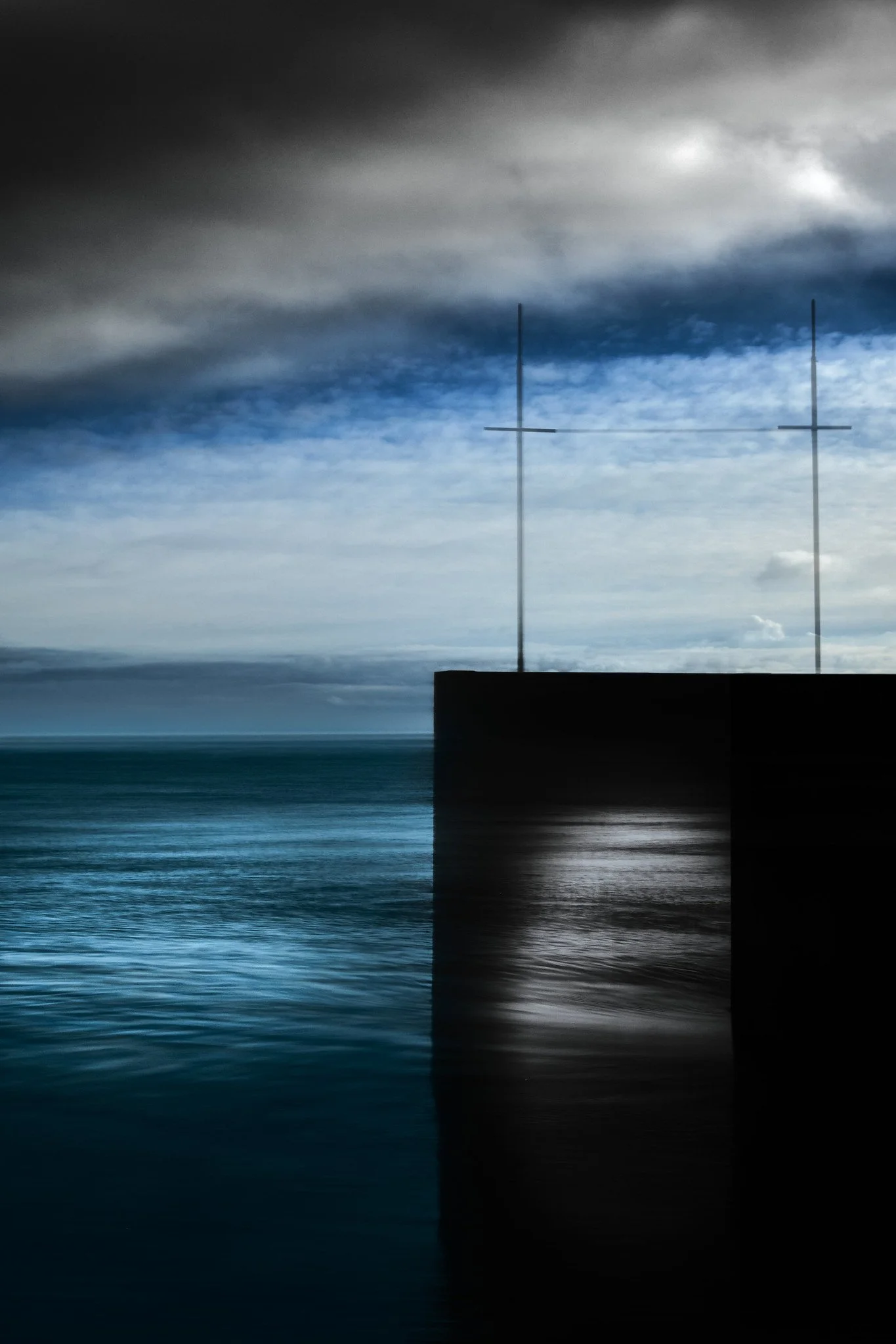 A dark, moody scene of a body of water with a concrete platform and two tall, thin structures with crossbars, set against a cloudy sky with areas of blue and gray.