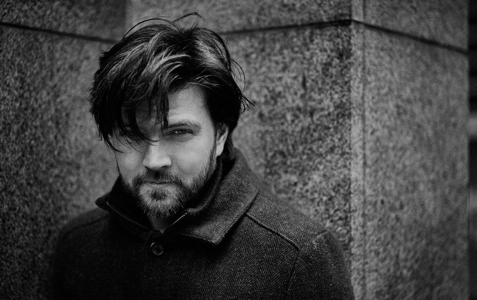 Black and white photo of a man with messy hair and a beard, leaning against a textured stone wall, looking into the camera.