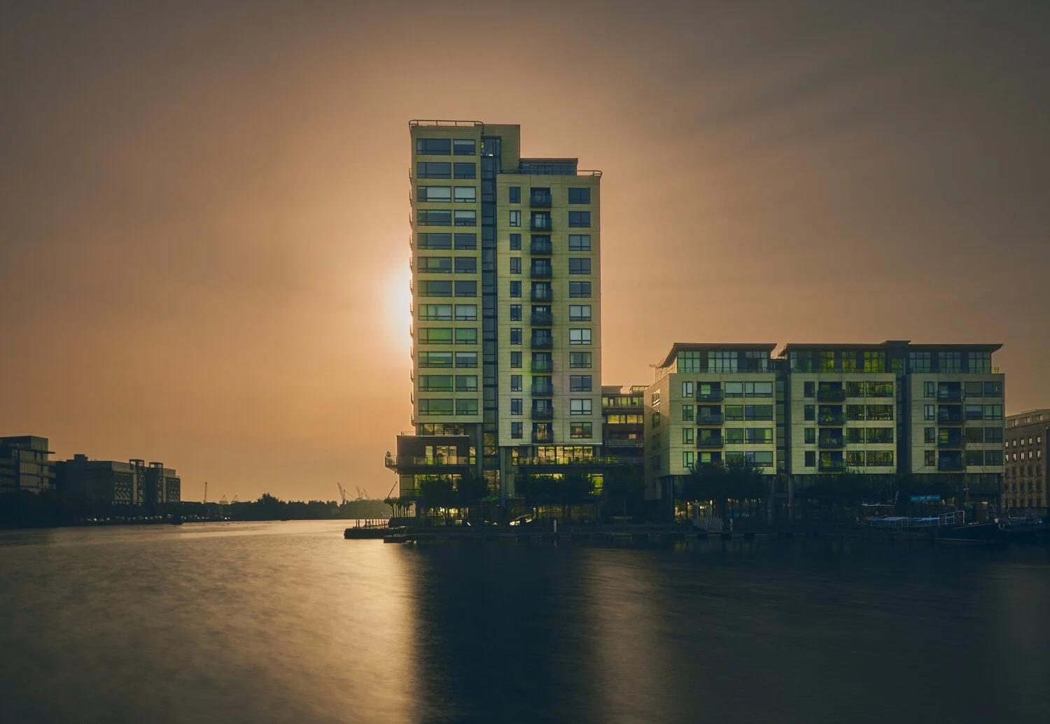 Modern high-rise buildings along a waterfront at sunset or sunrise, casting reflections on the water with a warm, hazy sky.