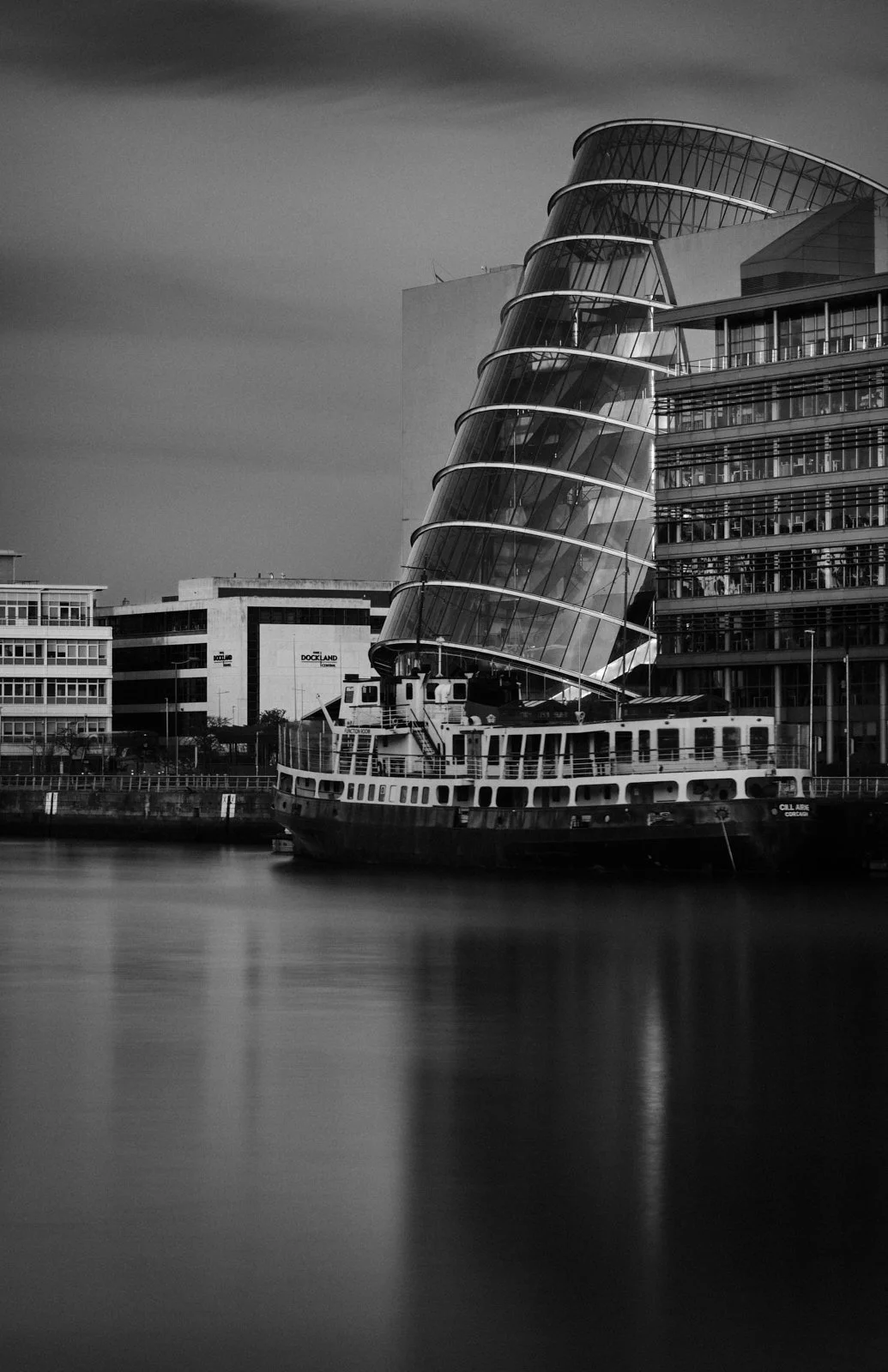 Black and white photo of a modern building with curved glass architecture and a boat docked along the water front.