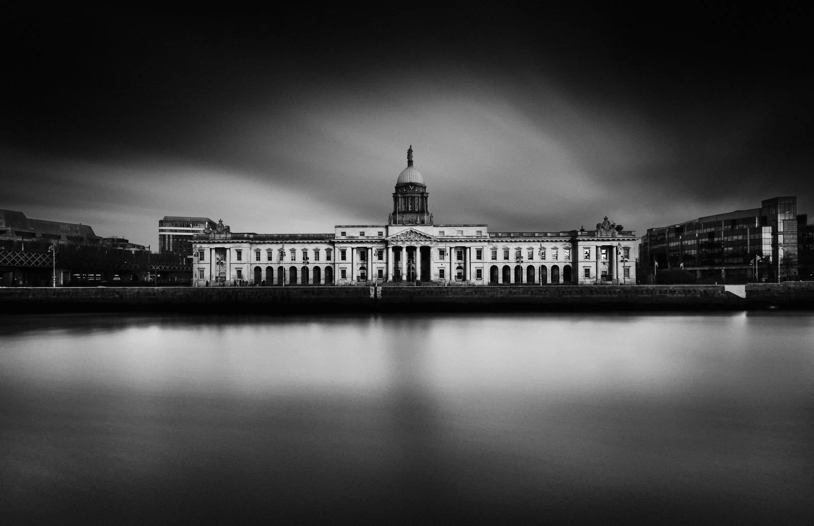 Black and white image of a historic government building with a dome, situated along a river, with modern buildings outside.