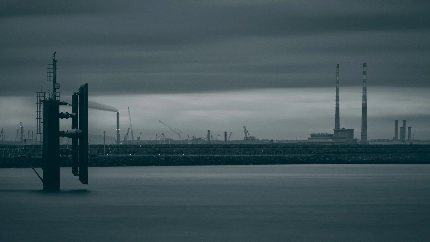 Industrial harbor scene with factories, smokestacks, and cranes under a cloudy sky, viewed over water.