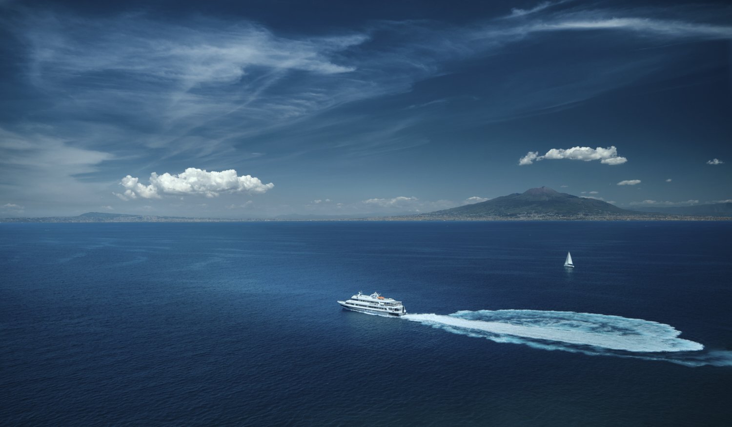 A large white boat moving fast across a deep blue ocean, leaving a foamy wake behind. In the distance, a sailboat navigates the water near a mountainous island under a partly cloudy sky.