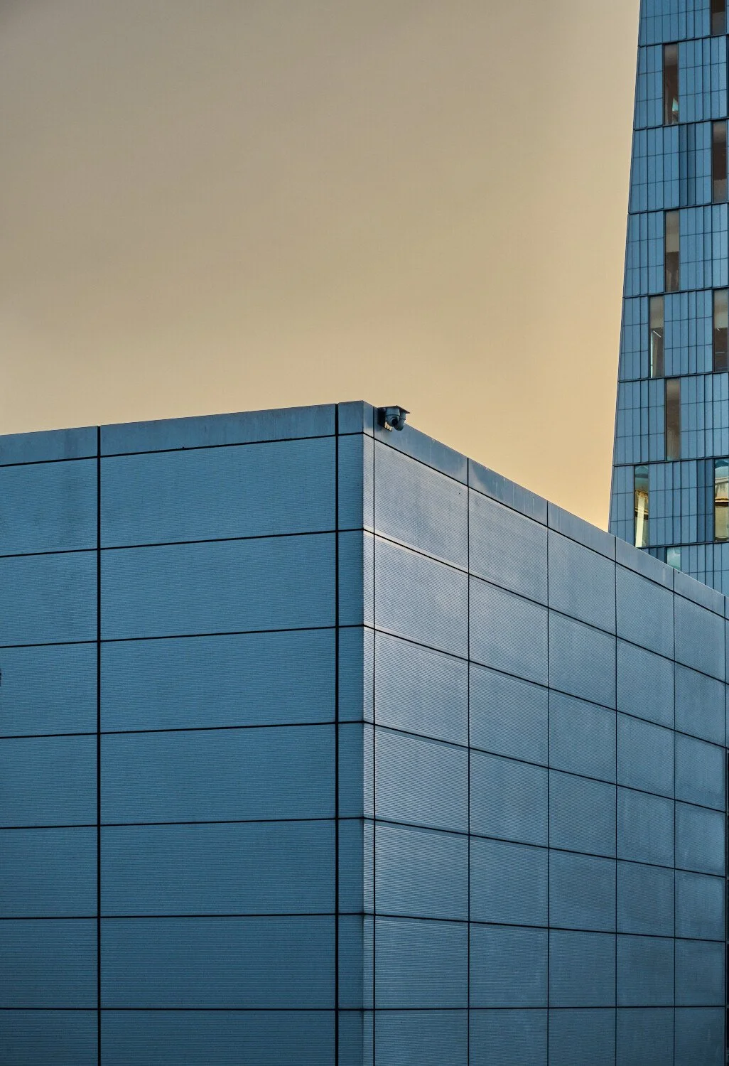 Close-up of modern blue building with a security camera mounted on the corner, against a beige sky and another tall glass building in the background.